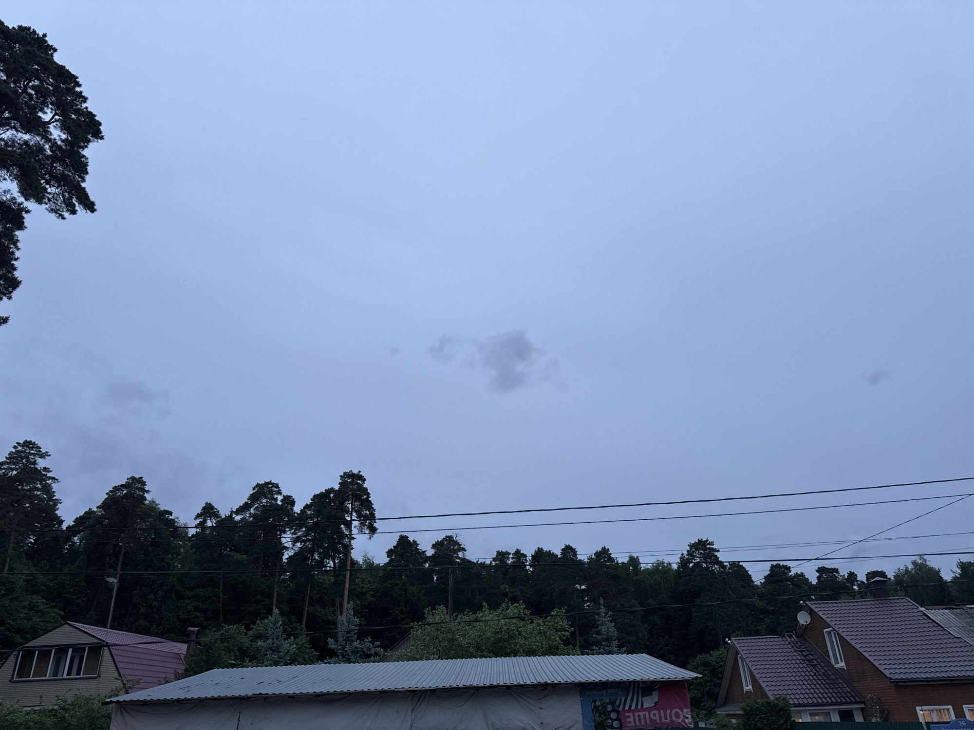 The image depicts a landscape in what appears to be Balashikha, Russia. The sky is overcast with a few wispy clouds. Tree lines are visible. A power line runs horizontally across the mid-ground. Below the power lines, rooftops of buildings are discernible, including a corrugated metal roof, and a brown roof. The image composition is static. No people or specific interactions are evident.