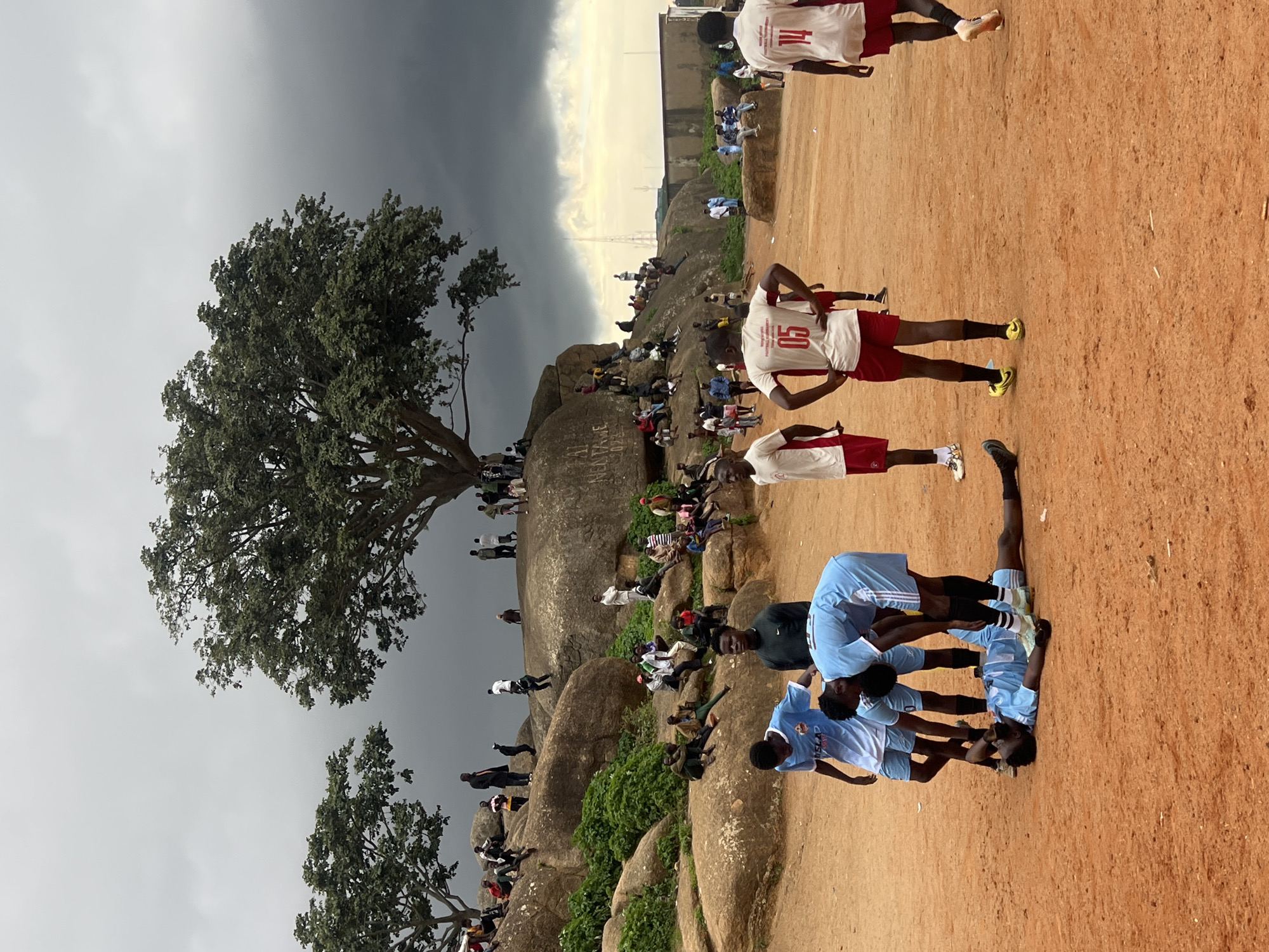 The image depicts a group of young men in athletic attire, some in white jerseys with red shorts and others in light blue jerseys with dark shorts, gathered on a dusty, reddish-brown field. The scene is set against a backdrop of large, weathered rocks and lush green foliage, suggesting a natural, outdoor environment. A large, leafy tree with a thick trunk stands prominently on the left side of the frame, its branches reaching towards a dramatic sky.
The individuals appear to be engaged in a sporting event, possibly a soccer match or practice, as indicated by their uniforms and the presence of a cleared area resembling a field. Some are standing, while others are in various poses of physical activity or rest. The time of day is uncertain, but the lighting suggests it could be late afternoon or early evening, with a mix of diffused sunlight and ominous, dark clouds gathering overhead, hinting at a change in weather. The emotions conveyed are difficult to discern, but the focus and camaraderie among the athletes suggest a shared purpose.
Notable details include the weathered texture of the rocks, the dense greenery, and the contrast between the dry earth and the overcast sky. There is some visible text on the jerseys, though it is partially obscured or difficult to read clearly. On one white jersey, the number "05" is visible, and on another, "14". The location context of Jos, Nigeria, evokes a specific geographic and cultural setting. The overall impression is one of vibrant activity within a rugged, natural landscape, underscored by a dynamic sky.