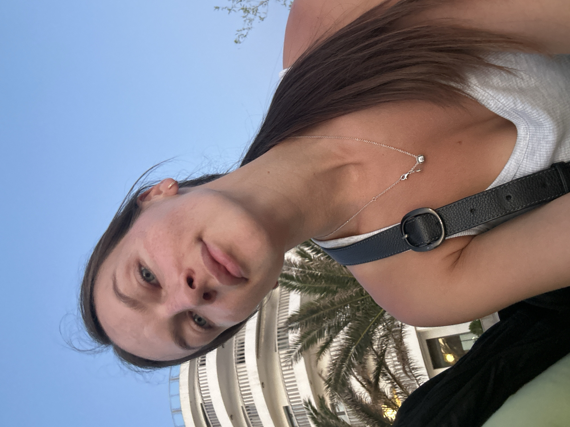 A woman with brown hair in a ponytail takes a selfie outdoors with a modern, white building and palm trees in the background, suggesting a location in the city of Bečići, Montenegro. The sky is clear and blue. She is wearing a white tank top and a silver necklace. A black purse strap with a buckle is visible across her chest.