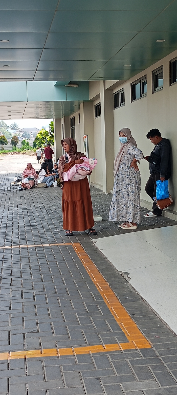 The scene is an outdoor area in Soreang, Indonesia, located at coordinates -7.032 latitude and 107.527 longitude.  The image shows several people standing and sitting on a brick pathway under an overhang with a light blue ceiling. There are three women. The first is wearing a brown dress and a head scarf, and is holding a baby. She is facing forward.  The second woman is wearing a white and floral dress, a head scarf, and a blue face mask. She is facing forward with her right hand on her hip.  The third woman is wearing a blue head scarf and a blue jacket. She is sitting on the ground with her legs crossed and is looking down. There is a man in a black hooded sweatshirt with a blue bag slung over his shoulder, and a blue mask around his neck. He is facing forward with his back to the wall and his right hand on a purse that is hanging on the wall. Two other men are standing farther back from the group. The pathway is made of gray bricks and has yellow lines painted on it. There is a curb between the brick pathway and the wall. There is a row of windows above the curb. The sky is visible. The ground is covered in grass and plants.