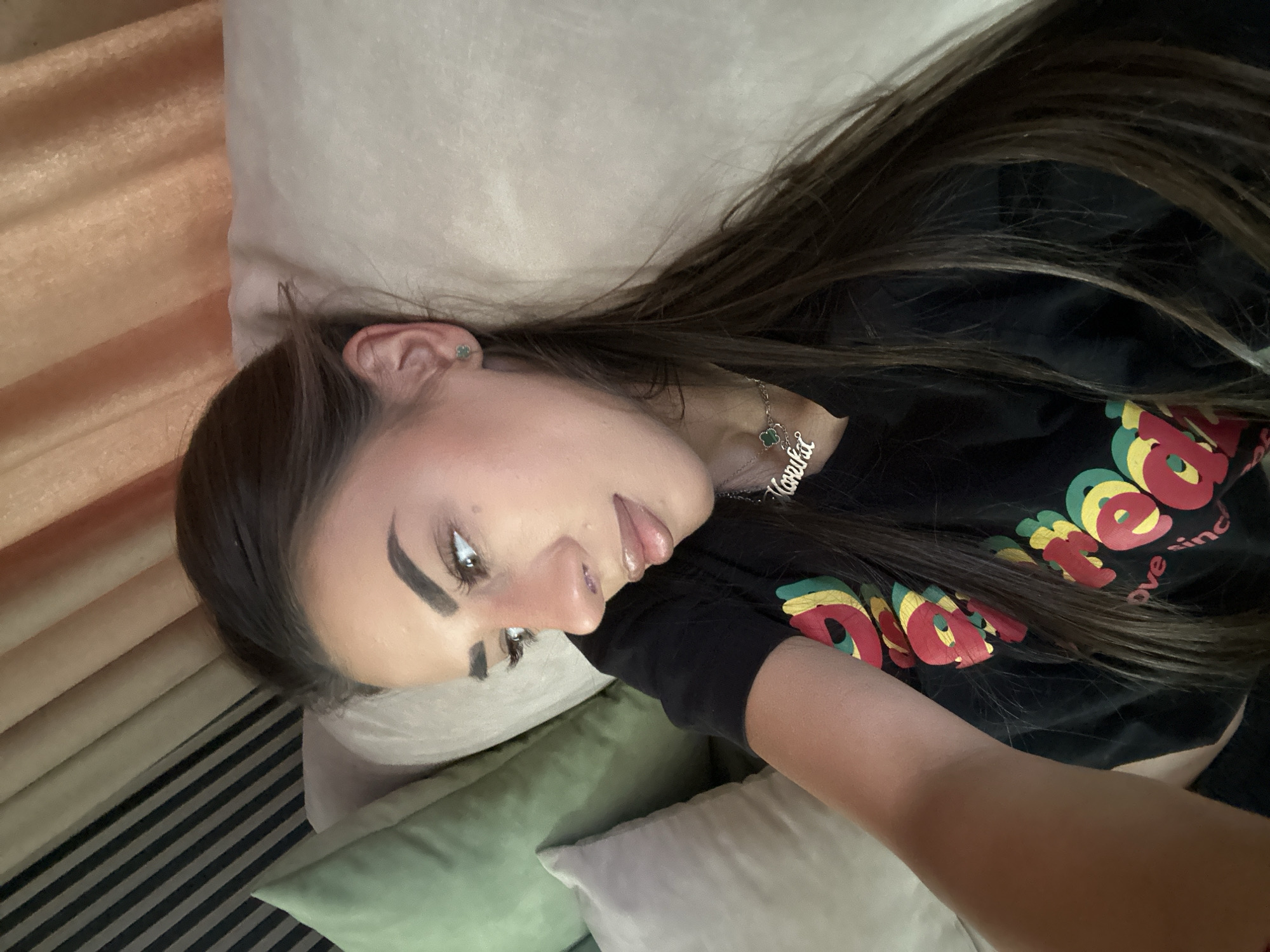 In Sofia, Bulgaria, a woman is lying down on a couch, with a pillow under her head and another pillow behind her. She is wearing a black t-shirt with a colorful design on it and a silver necklace with her name on it. She has long brown hair and is wearing a small green earring. The background of the image consists of a striped curtain and a textured wall.