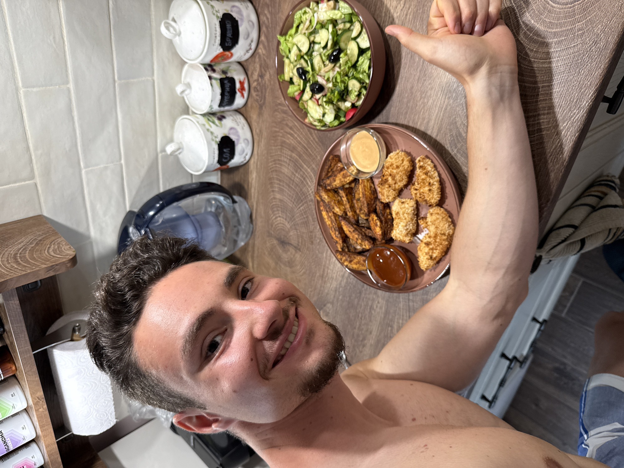 A young adult male, shirtless with short dark hair and light stubble, smiles directly at the camera while giving a "thumbs up" gesture with his right hand. The scene is an indoor kitchen in Sofia, Bulgaria. On a patterned wooden countertop in the foreground, a meal is displayed across two dark terracotta-colored dishes. One plate holds golden-brown breaded chicken nuggets, a pile of dark seasoned potato wedges, and two small glass ramekins containing a reddish-brown sauce and a lighter creamy yellow-orange sauce. Adjacent to this, a bowl contains a fresh green salad with chopped lettuce, sliced cucumbers, red radishes, and black olives. In the background, three white ceramic spice jars with lids are visible, featuring black labels with white Cyrillic text identifying them as "СОЛ" (Salt), "ЧУБРИЦА" (Savory), and "БРАШНО" (Flour). A light-colored subway tile backsplash spans the wall behind the jars. To the left, a dark-colored electric kettle is partially visible, and a white paper towel roll on a holder hangs from a wooden shelf above it.