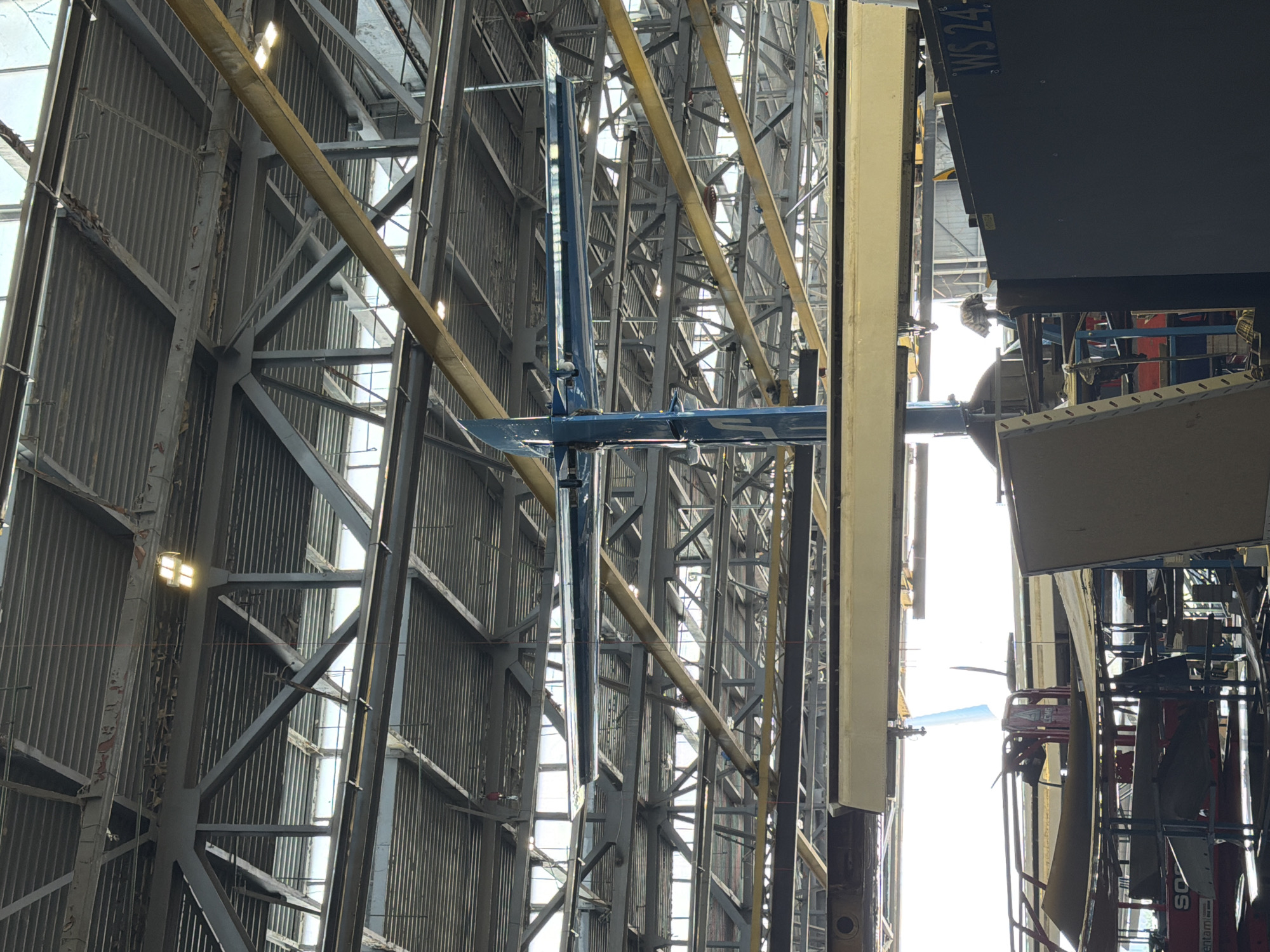 This media file captures the interior of a large industrial building, likely an aircraft hangar or a specialized manufacturing facility, located in Sofia, Bulgaria. The perspective is looking upwards and across the vast space, dominated by robust metal structural elements.
The scene is characterized by a complex lattice of grey steel beams and trusses forming the framework of the building's walls and ceiling. Many of these beams run vertically and diagonally, supporting corrugated metal wall panels. Bright rectangular LED lights are visible, mounted on the structure, providing illumination. Natural light also floods in from windows or translucent panels higher up, creating bright streaks against the darker structural elements. Several prominent yellow horizontal beams cross the frame, adding a contrasting color to the grey metalwork.
In the central and right portions of the image, large industrial objects are visible. A striking blue structure, resembling the vertical stabilizer and a part of the horizontal stabilizer (tail assembly) of an aircraft or a large drone, stands out. This sleek, fin-like component has a darker blue leading edge and a lighter blue main body, with subtle details suggesting its aerodynamic purpose. To the right of this blue component, a large dark grey or black flat surface is seen, possibly part of the fuselage or another major section of a vehicle. On this dark surface, the text "WS 24-" is partially visible, hinting at an identification number or model designation.
Further to the right, other industrial equipment, including what appears to be scaffolding or a mobile work platform with a red base, and various tools and materials, are arranged. No people are visible in the image, suggesting the focus is on the scale of the facility and the objects within it. The overall impression is one of an active industrial environment dedicated to the construction, assembly, or maintenance of large-scale mechanical components, likely aeronautical. The bright lighting suggests it is daytime.