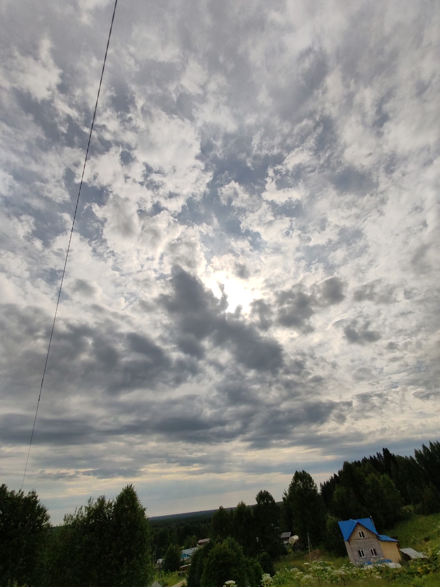 A wide-angle, high-angle shot captures a cloudy sky with the sun peeking through the clouds on the right side of the frame. A thin black wire extends diagonally from the top left to the bottom left of the image. Below the sky, a dense line of green trees stretches across the middle ground. On the right side of the frame, a small house with a bright blue roof and a beige exterior is visible nestled among the trees. The house has several windows. In the foreground, there are green trees and some white wildflowers. The landscape appears to be rural. This image was taken in Chukhlem, Russia.