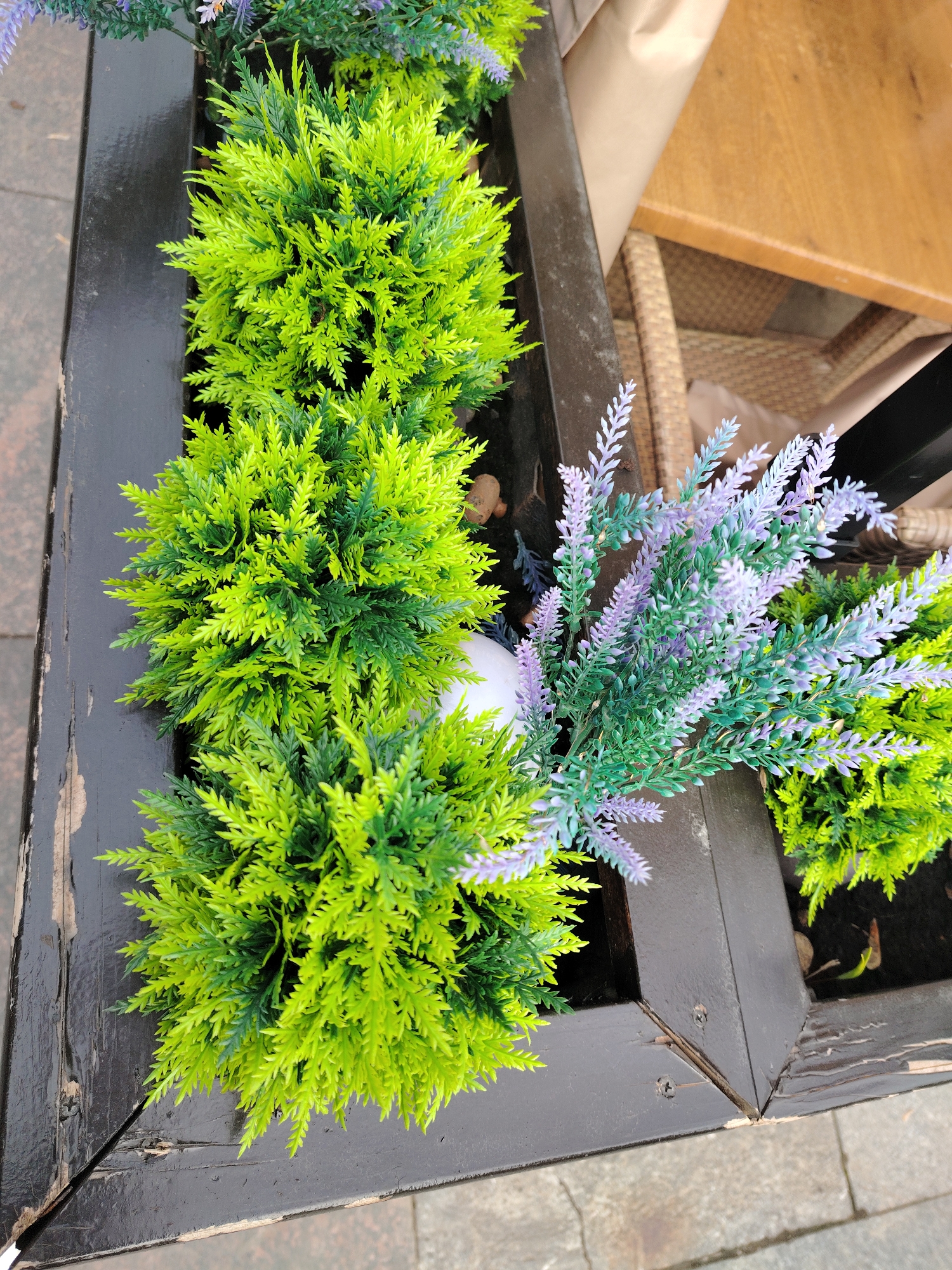 This is an overhead, close-up shot of a dark wooden planter box filled with artificial plants, situated outdoors in Moscow, Russia. The planter is made of dark brown or black painted wood, showing signs of wear with chipped paint and visible wood grain, especially on the top edge and at a corner joint secured by screws.
The planter holds several clusters of artificial foliage. Dominating the scene are bright lime green, shrub-like plants with dense, spiky leaves. Some areas of these plants exhibit a slightly darker green hue. Interspersed with the green plants are artificial lavender-like stems with small, light purple and light green blossoms. A small, round, white object is partially visible among the plants near the center of the planter.
To the left of the planter, the ground is paved with grey, rectangular stones. In the background, on the right side, parts of an outdoor seating area are visible. This includes a section of a light brown wooden table and a chair with a woven or wicker texture. A piece of light brown paper or fabric is also partially seen behind the table. The lighting suggests it is daytime. There is no visible text or specific activity occurring.