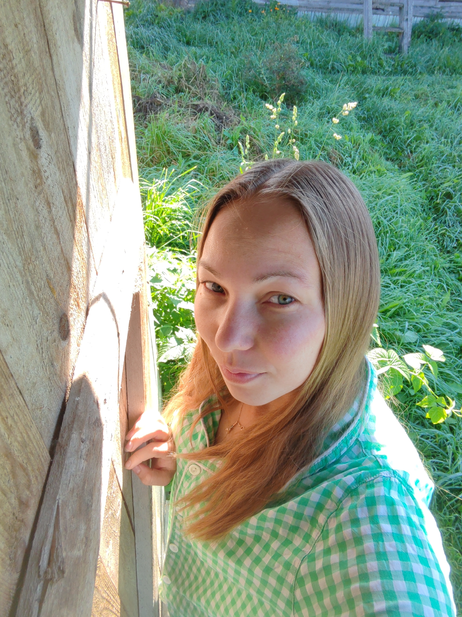 A young woman with blonde hair and green eyes is taking a selfie outdoors, leaning against a wooden structure. She is wearing a green and white checkered shirt and a necklace. The background consists of lush green grass and some foliage, suggesting a rural or garden setting. The lighting indicates it is daytime, with sunlight casting shadows on the wooden surface. The location is identified as Chukhlem, Russia.