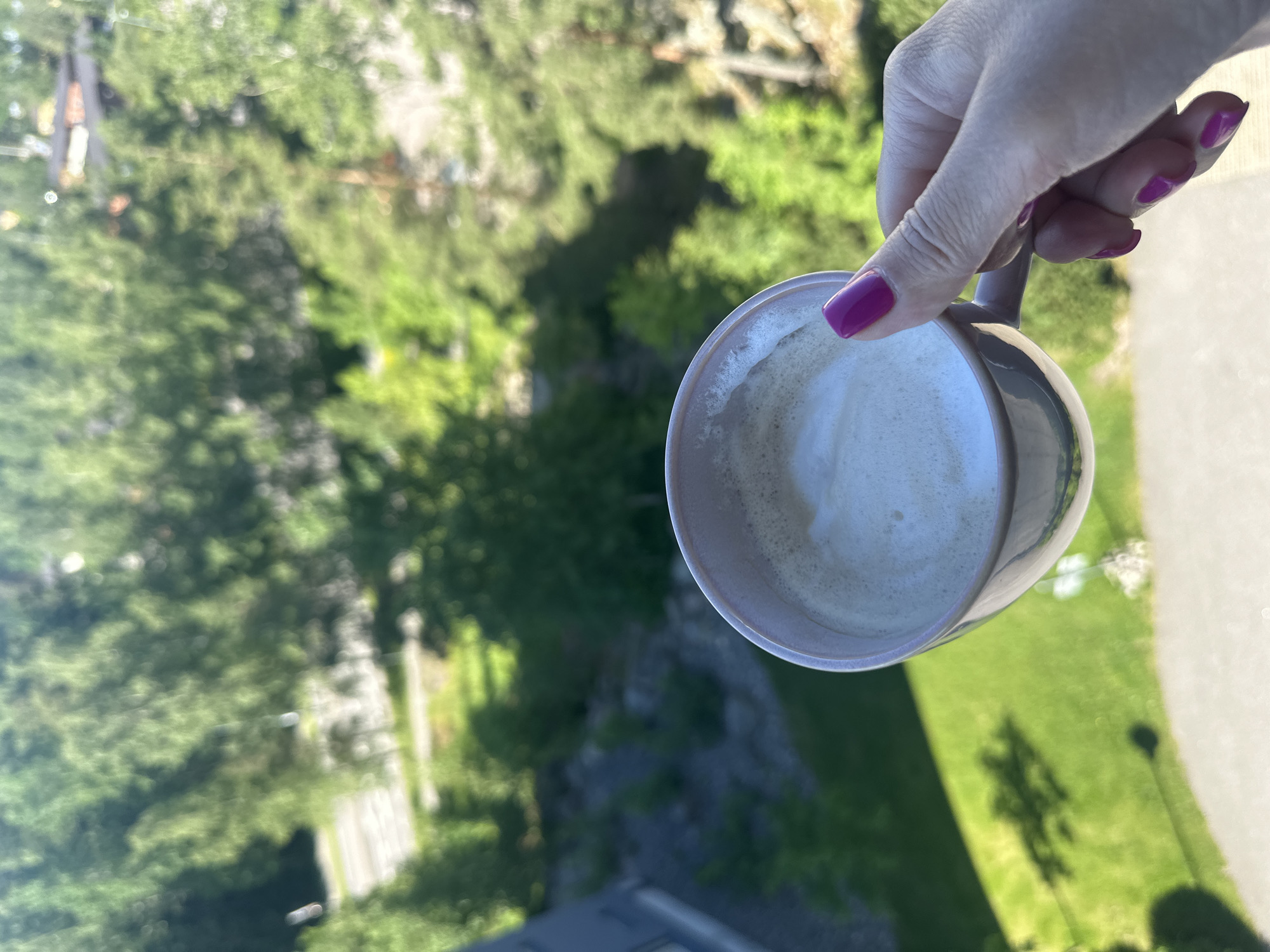 A close-up shot captures a woman's hand holding a light pink mug filled with coffee and foam. Her fingernails are painted a vibrant magenta. The mug is positioned in the foreground, with the background blurred by the shallow depth of field. The background shows a view of lush green trees and vegetation, suggesting an outdoor setting, possibly a balcony or window overlooking a natural area. The lighting indicates it is daytime, likely morning or afternoon, with sunlight casting shadows.