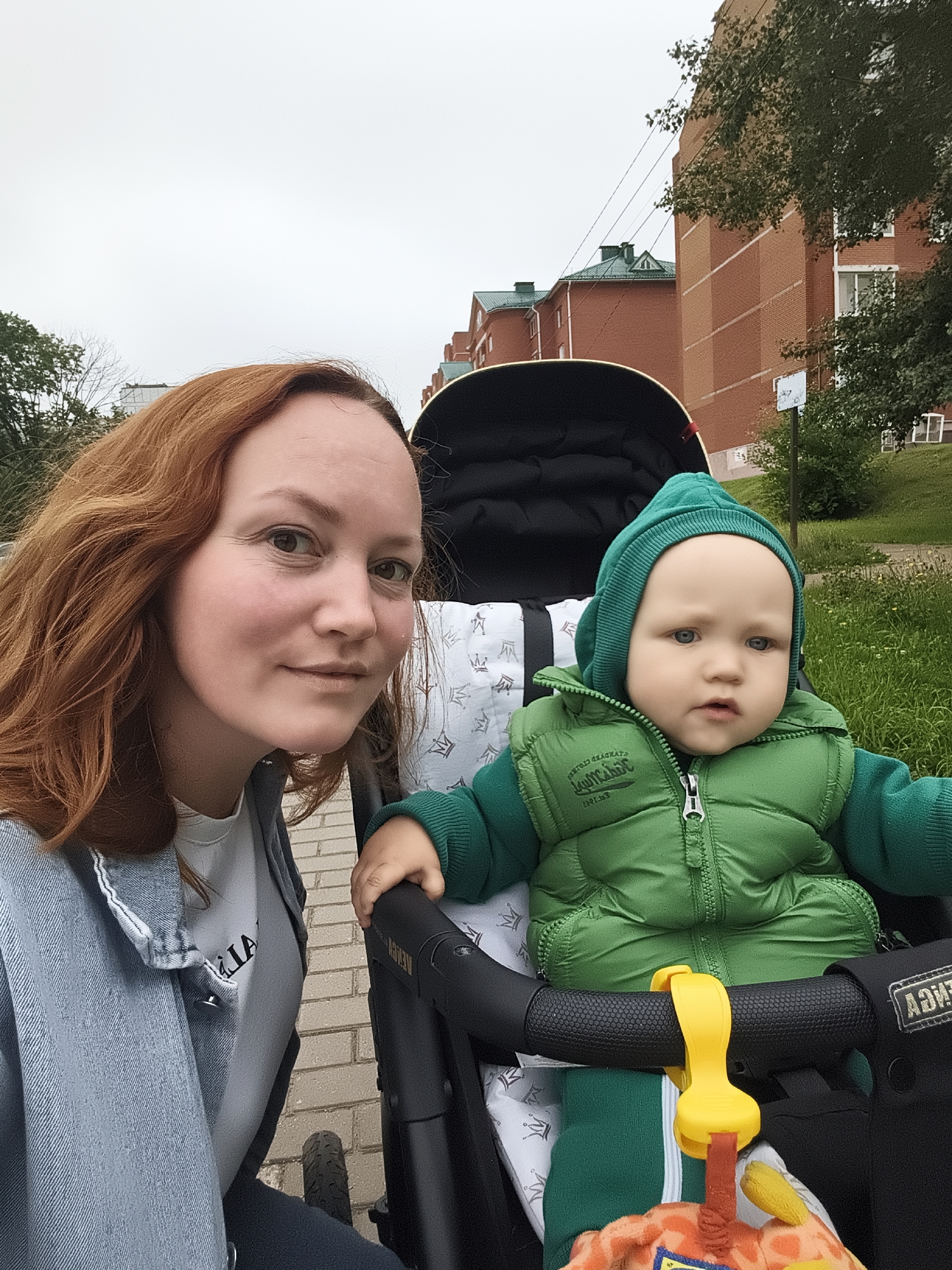 A woman with reddish-brown hair and a light-skinned complexion, wearing a light blue denim jacket over a white t-shirt, is positioned on the left side of the frame, looking towards the camera with a slight smile. To her right, a baby is seated in a stroller, wearing a green hooded jacket and a green quilted vest. The baby has fair skin and light blue eyes, and is looking slightly off-camera. The stroller has a black frame and a black canopy. A yellow clip is attached to the stroller handle. The background shows residential buildings with red brick facades and green roofs, characteristic of Chaikovsky, Russia. There is also greenery and a paved walkway. The sky is overcast.