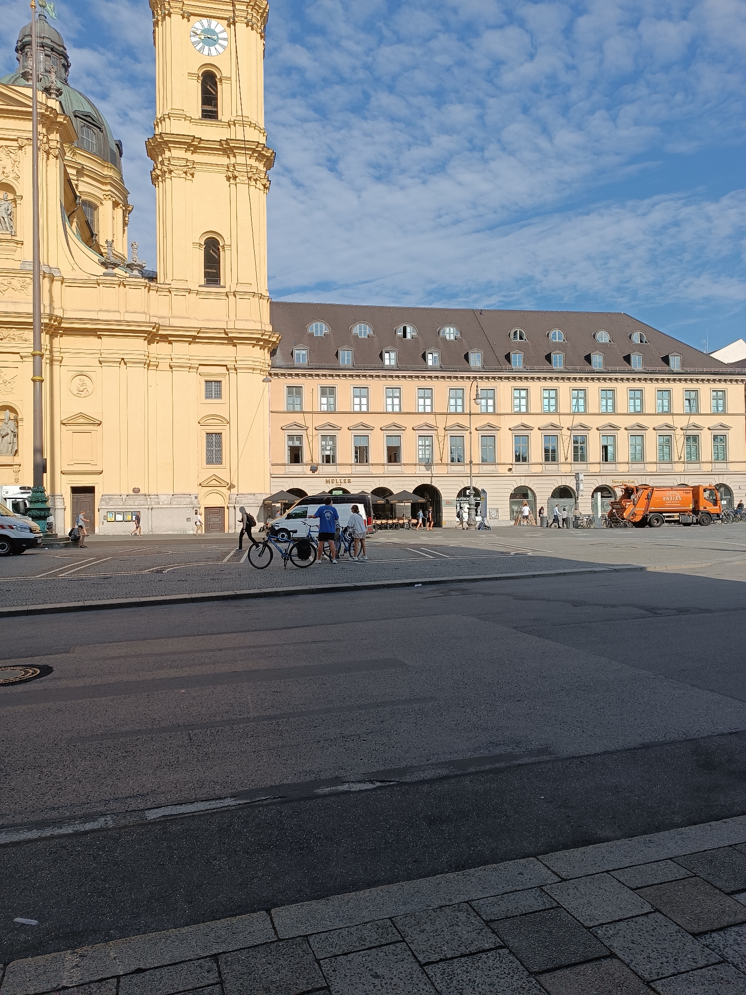 The image depicts a public square in Munich, Germany, likely Marienplatz, dominated by the yellow facade of a historic building, possibly a church or Rathaus, with a tall clock tower. In front of this building, a beige, multi-story structure with arched ground-floor entrances and numerous windows runs horizontally. The sky above is blue with scattered white clouds.
In the foreground, a paved area transitions to asphalt. Several people are present in the square. A group of three individuals are standing near a white van and two bicycles. Another person walks towards the building on the left, and several more figures are visible near the arched entrances of the beige building, suggesting shops or cafes. A bright orange garbage truck is parked on the right side of the square, with the word "MULLER" and "ENZIAN" visible on its side. Bicycles are also parked near this truck. The overall atmosphere suggests a sunny day in a bustling urban environment.