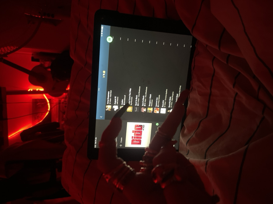A tablet screen displays a YouTube Music playlist, with song titles like "Army Dreamers," "August 10," "Avalon," "by Love Your Way," and "Back To Back" visible. A "Create" button sits above a large album cover that says "Dido". A pair of hands with long fingernails and multiple rings rests on the tablet. The scene is bathed in red light, illuminating bedding with thin white stripes, and a guitar and fan are visible in the background.