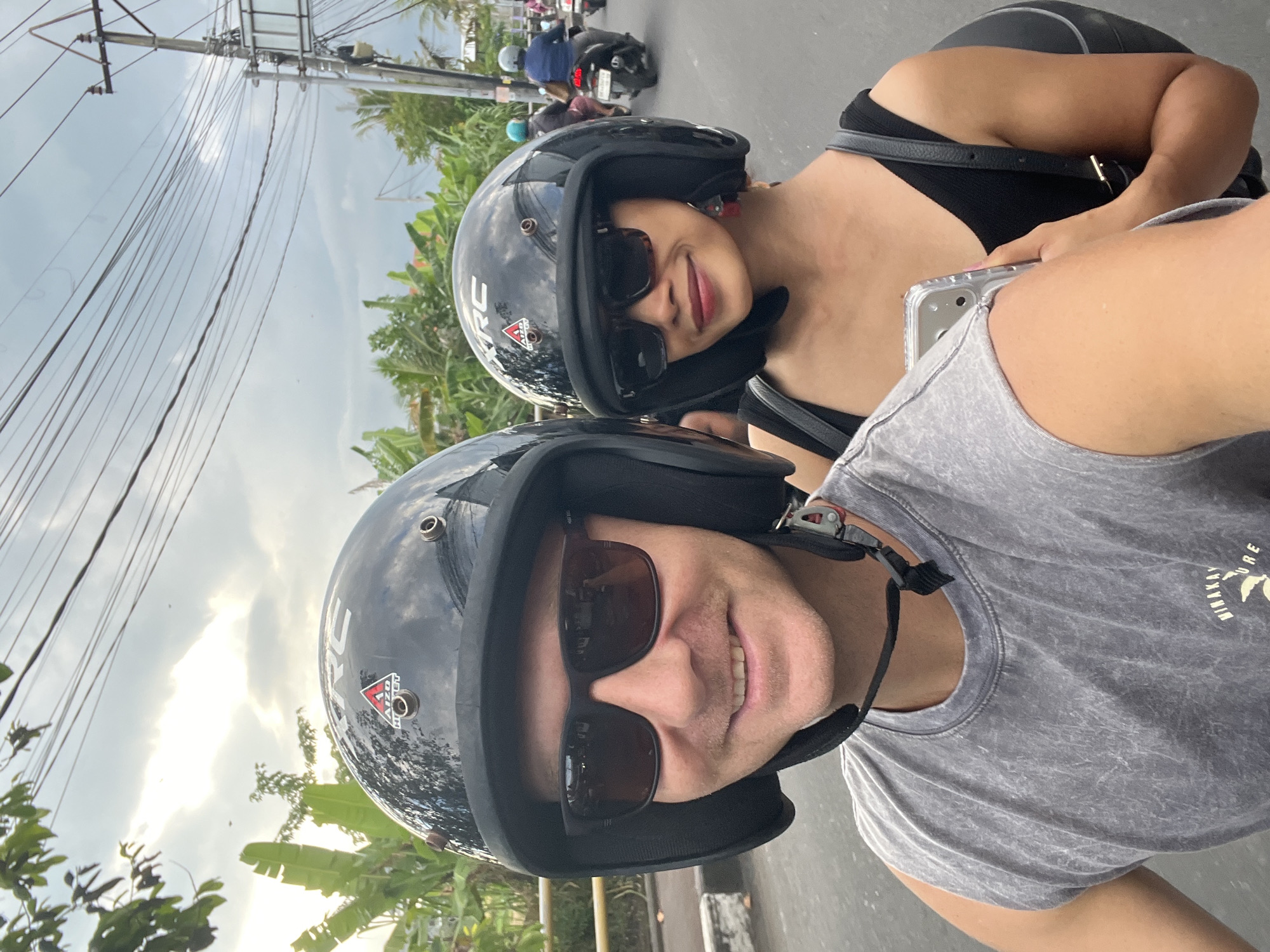 A medium shot, rotated counterclockwise, shows two people riding a scooter in North Kuta, Indonesia. The foreground features a person wearing sunglasses and a helmet, a gray t-shirt with "DINAKAY NATURE" printed on it, and a black strap. Adjacent to them is another person, also wearing sunglasses and a helmet, dressed in a black top with a strap. A cell phone is visible in the hands of the first person, suggesting they are taking the photograph.
The background includes other scooters on the road, lush green trees and plants, and utility poles with wires. The sky is overcast. The scene suggests the individuals are engaged in casual travel or sightseeing.