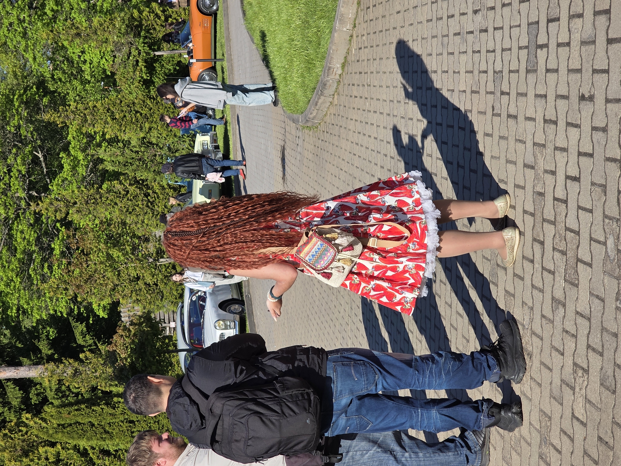 The image shows a sunny outdoor scene in Repino, Russia, where a group of people are viewing classic cars. In the foreground, a woman with long braided red hair, wearing a red and white patterned dress with white lace trim, tan stockings, and gold shoes, stands on a paved walkway. She has a small, colorful bag on her shoulder. A man wearing jeans, a black jacket, and a backpack stands next to her. The paved walkway is bordered by a grassy area. Further in the background, various classic cars of different colors are visible, along with more people standing and looking at the cars. The presence of trees suggests the area might be near a park or a wooded area. The shadows suggest a sunny day.