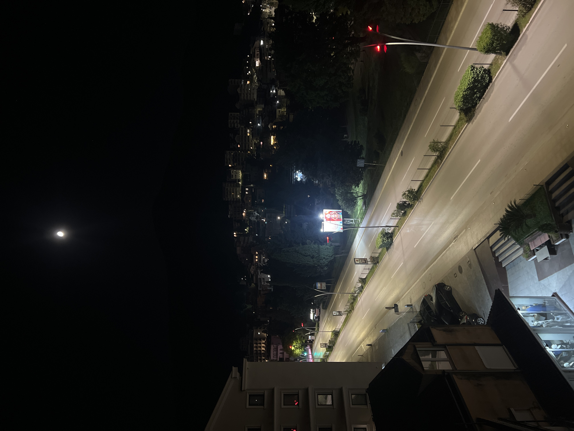 The image is taken at night in Budva, Montenegro, showing a view from a high vantage point, possibly a building. A full moon is visible in the dark sky. The scene includes a well-lit street running through the city. There are buildings along the street with illuminated windows, and the street lighting highlights the road surface. A traffic light is visible at the intersection. Along the side of the road are plants and bushes. There is parked vehicle along the building. There is also a billboard along the street. The image captures a combination of urban and natural elements in the nighttime environment of Budva.