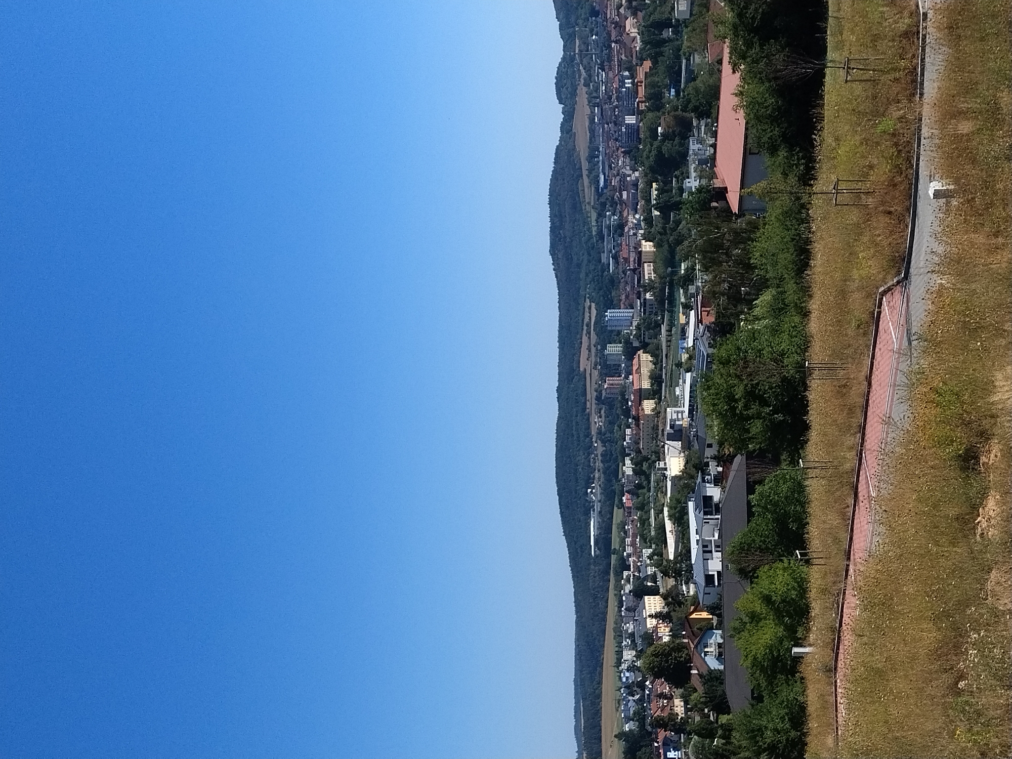 A wide, high-angle shot shows a clear blue sky above a sprawling cityscape nestled in a valley. The city, identified as Písek in the Czech Republic, is characterized by a dense collection of buildings of varying heights and architectural styles, interspersed with patches of green trees. In the foreground, a grassy, overgrown hillside slopes down towards a paved area with a red-bricked border. Several young trees, staked to remain upright, are sparsely planted on the hillside. The background features rolling hills covered in dark green trees, suggesting a rural or semi-rural environment surrounding the urban area. The overall scene depicts a sunny day with good visibility.