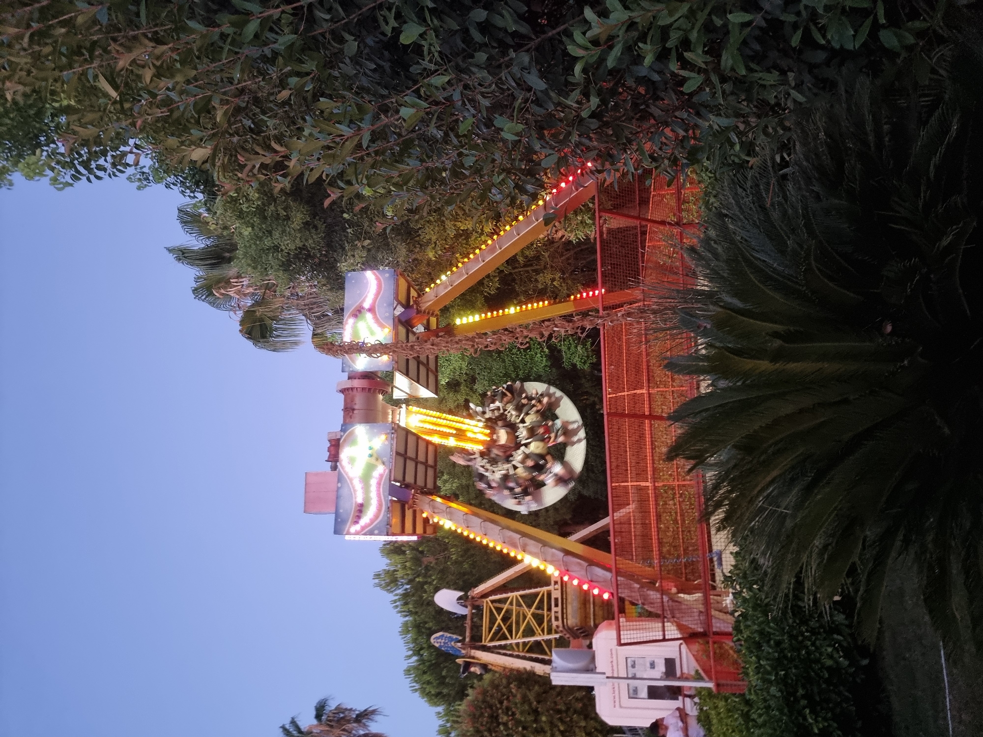 This is a medium-shot, eye-level, vertical photograph of an amusement park ride in Manavgat, Turkey. The ride, a swinging pendulum attraction, is centrally located and in motion, with a circular gondola filled with riders spinning. The ride is brightly lit with yellow and red lights along its support beams, and the top of the structure has a colorful, illuminated sign. To the left of the main ride, a yellow metal structure with a crane-like element is visible, and a white booth with a person standing in front of it is partially obscured by the structure. The background consists of lush green trees, including palm trees, and a clear, light blue sky. In the foreground, a large, dark green palm-like plant with spiky fronds is prominent. The overall impression is of a lively, evening scene at an amusement park.
