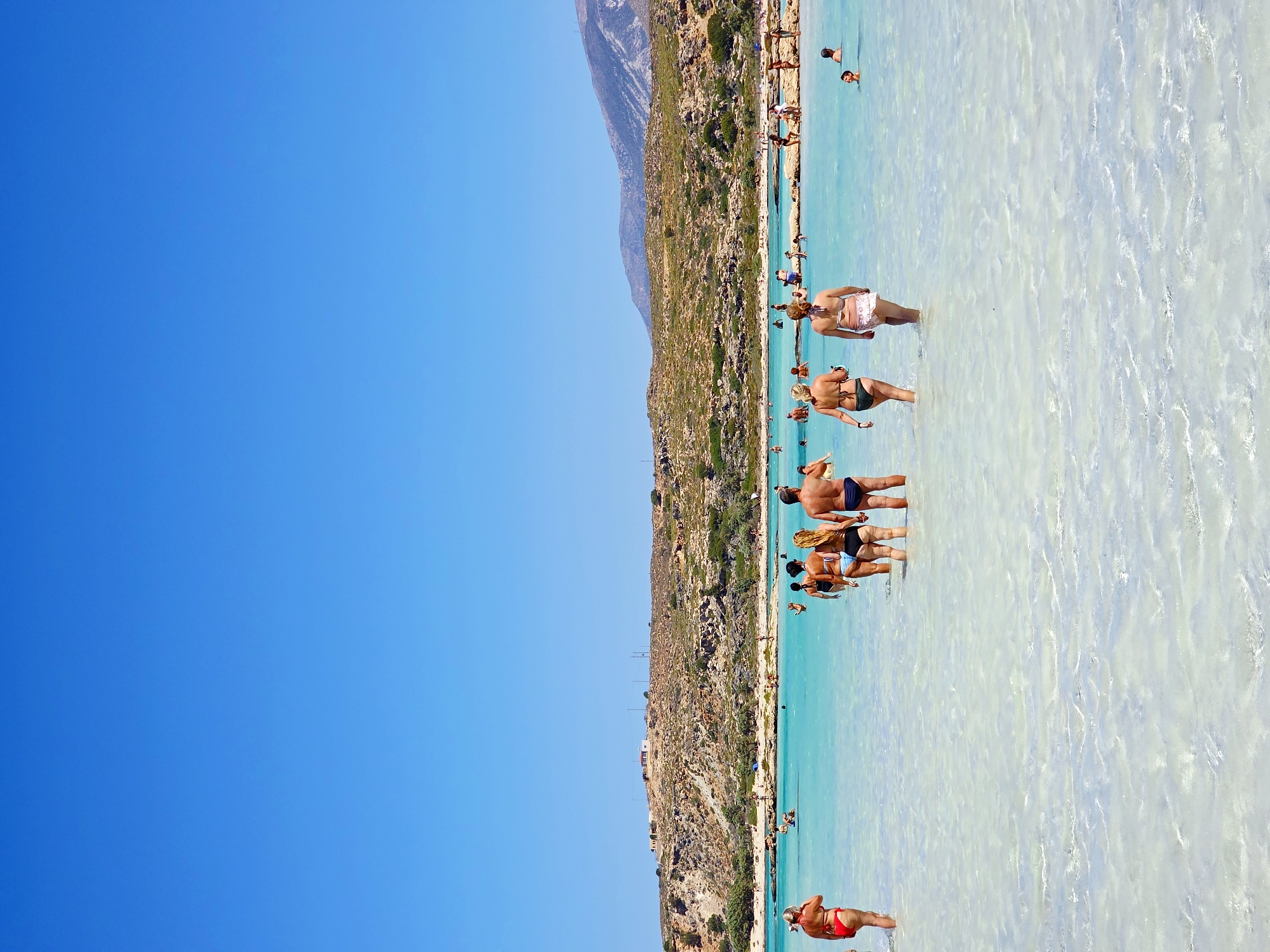 The image depicts a clear, sunny day at a beach with turquoise water and a sandy bottom. A group of people are wading in the shallow water, some walking, others standing. In the foreground, a woman in a red bikini is facing away from the camera. To her right, a group of four people are walking towards the center of the frame. In the background, a rocky, arid hillside with sparse vegetation stretches towards the horizon, topped by a few scattered buildings and communication towers. The sky is a brilliant, cloudless blue. The scene suggests a popular tourist destination, likely Greece given the mention of "Unknown, Greece" in the prompt, though the specific location is not identifiable from the image itself.