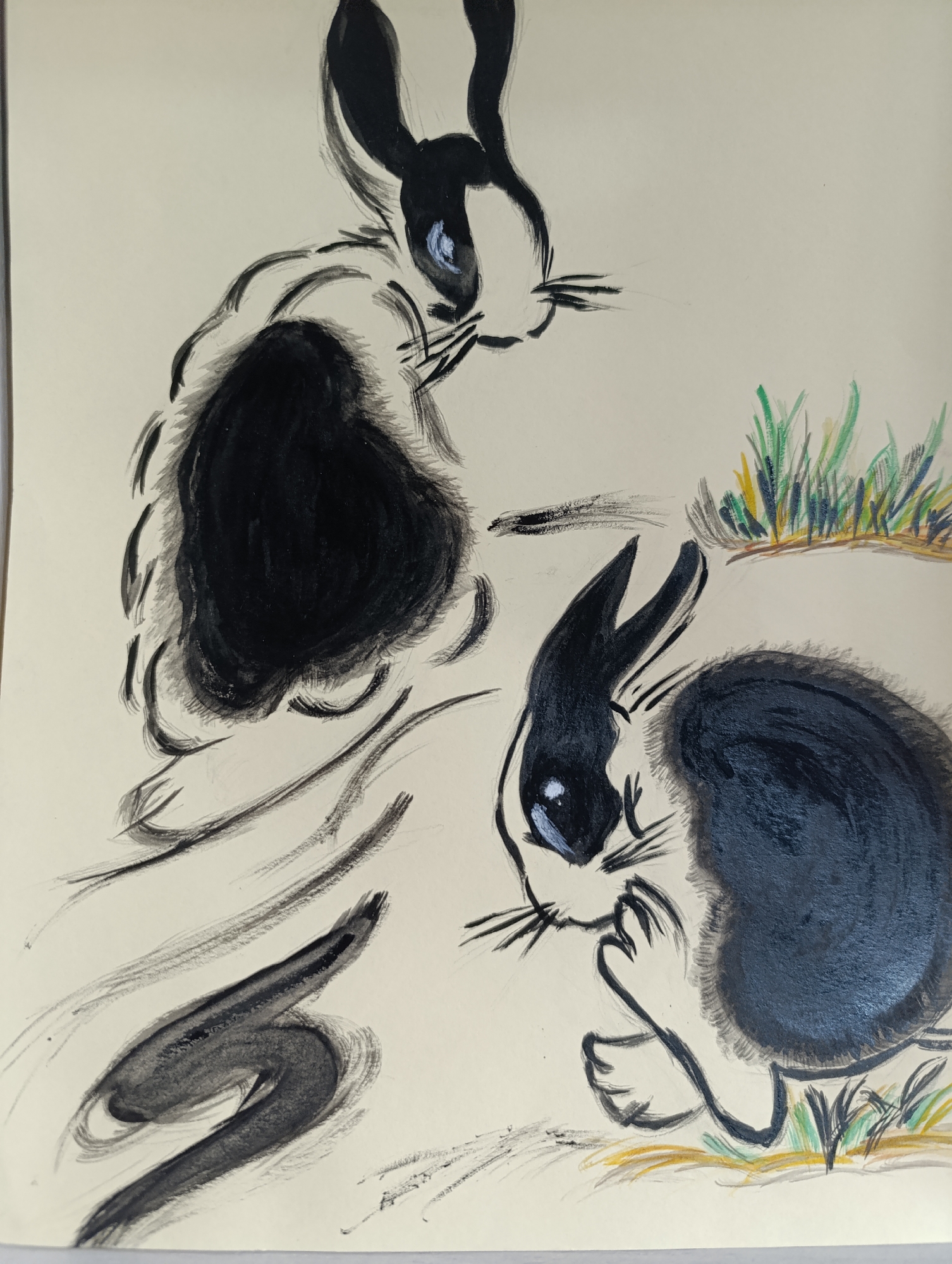 An expressive, somewhat abstract drawing featuring two black and white rabbits on a light-colored ground. The medium appears to be a combination of ink wash or watercolor for the black forms, with details in white, blue, and colored crayon/pastel for the grass.
The upper rabbit is positioned in the upper left quadrant, facing right. It has long black ears, a white face with blue eyes and white highlights, and distinct black whiskers. Its body is a large, dense black mass, with lighter black brushstrokes outlining its form, suggesting fur.
The lower rabbit is in the lower right quadrant, facing left. It also has black ears, a white face with blue eyes and white highlights, and black whiskers. One front paw is raised to its mouth or chin area, in a gesture often associated with grooming or contemplation. Its body is a rounded, solid black mass.
Between and beneath the two rabbits, and particularly extending from the upper rabbit towards the lower left, are several dynamic, abstract black brushstrokes. These vary in thickness and intensity, some suggesting movement, a tail, or ground texture, including a prominent S-shaped stroke in the lower left. In the upper right and beneath the lower rabbit, patches of grass are depicted with vertical strokes of green, yellow, and brown crayon or pastel. The overall scene is set in the city of Unknown, Unknown.