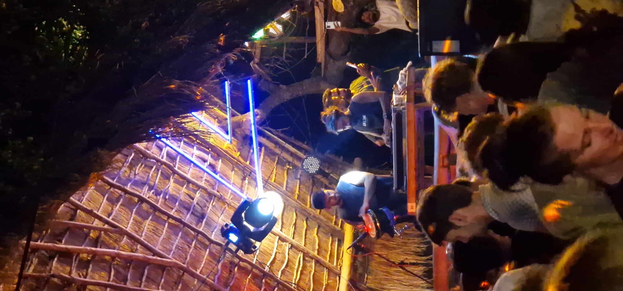 This image captures a nighttime musical performance in Pottuvil, Sri Lanka, within a semi-open-air venue. The roof structure is constructed from natural materials, likely thatch or dried palm leaves, supported by a framework of wooden beams. Bright blue LED strip lights are installed geometrically across these wooden beams, contrasting with the warm, ambient light illuminating the thatched roof.
On stage, a prominent white spotlight points downwards, illuminating the performance area, and a multi-LED PAR light is visible further back. Three male figures are engaged on the stage. To the left, a man with dark hair and a beard, wearing a dark sleeveless top, plays a hand drum mounted on a stand. In the center, another man, also with a beard and dark hair tied in a top knot, wears a dark sleeveless top with some white text, and operates electronic music equipment on a wooden table or console. Behind and slightly to the right of the central performer, a third man, with short dark hair and a beard, is visible holding a smartphone upright, seemingly recording or photographing the event.
The foreground is filled with the heads and upper torsos of a densely packed audience, observing the performance. In the background, to the far right, another individual in a white t-shirt holds a smartphone aloft, potentially recording. The overall scene depicts a lively concert atmosphere under artificial lighting, with elements of rustic construction.