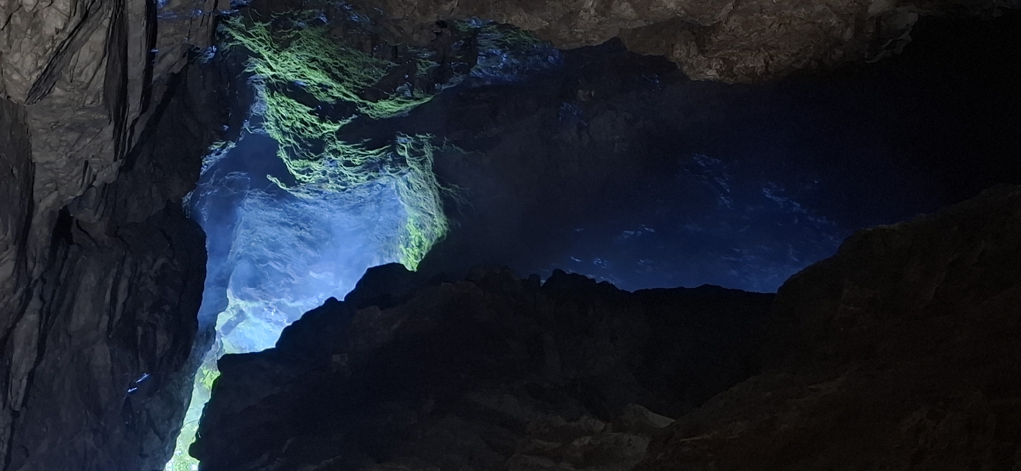 The image depicts the interior of a cave, likely the Devil's Throat cave in Trigrad, Bulgaria. The scene is characterized by dramatic rock formations and a striking contrast between light and shadow. A shaft of bright blue light streams in from an opening above, illuminating a misty, cavernous space. The rock walls are dark and textured, with patches of vibrant green moss clinging to some surfaces. The depth of the cave is emphasized by the diminishing light and the dark, shadowy depths below. The overall impression is one of natural grandeur and mystery, showcasing the geological features of the region.