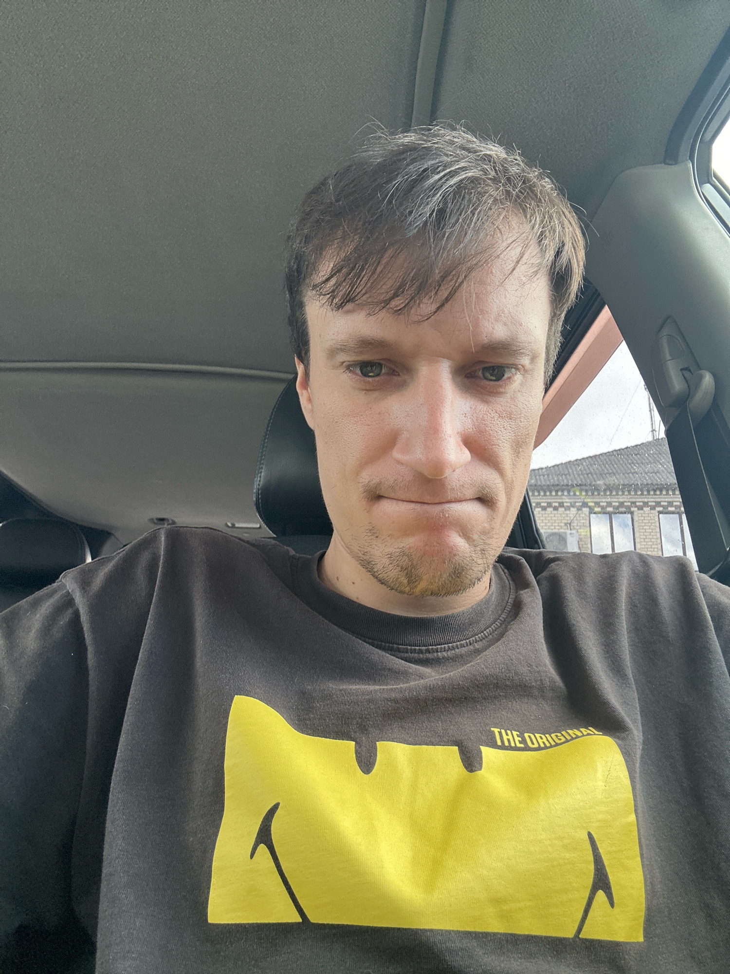 A man with light brown hair and a goatee, wearing a dark grey sweatshirt with a yellow smiley face graphic, is seated inside a car. The car's interior, including the ceiling and headrest, is visible. The man is looking down and slightly to the left with a neutral expression. Outside the car, a brick building with windows and an air conditioning unit is visible. The context of Shatava, Ukraine is not discernible from the image.
