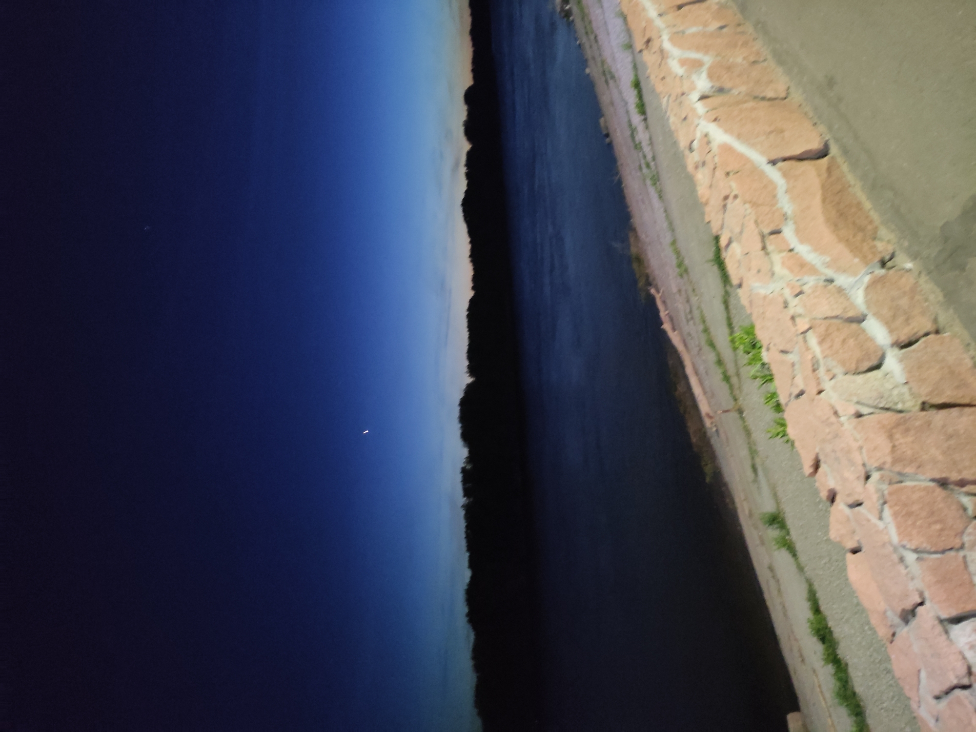The image depicts a night scene along the Irtysh River in Pavlodar, Kazakhstan. The camera faces east, capturing the darkening sky. A line of trees borders the river. A stone embankment runs along the riverbank. A crescent moon is visible in the upper-left portion of the frame. There are no people present in the scene.