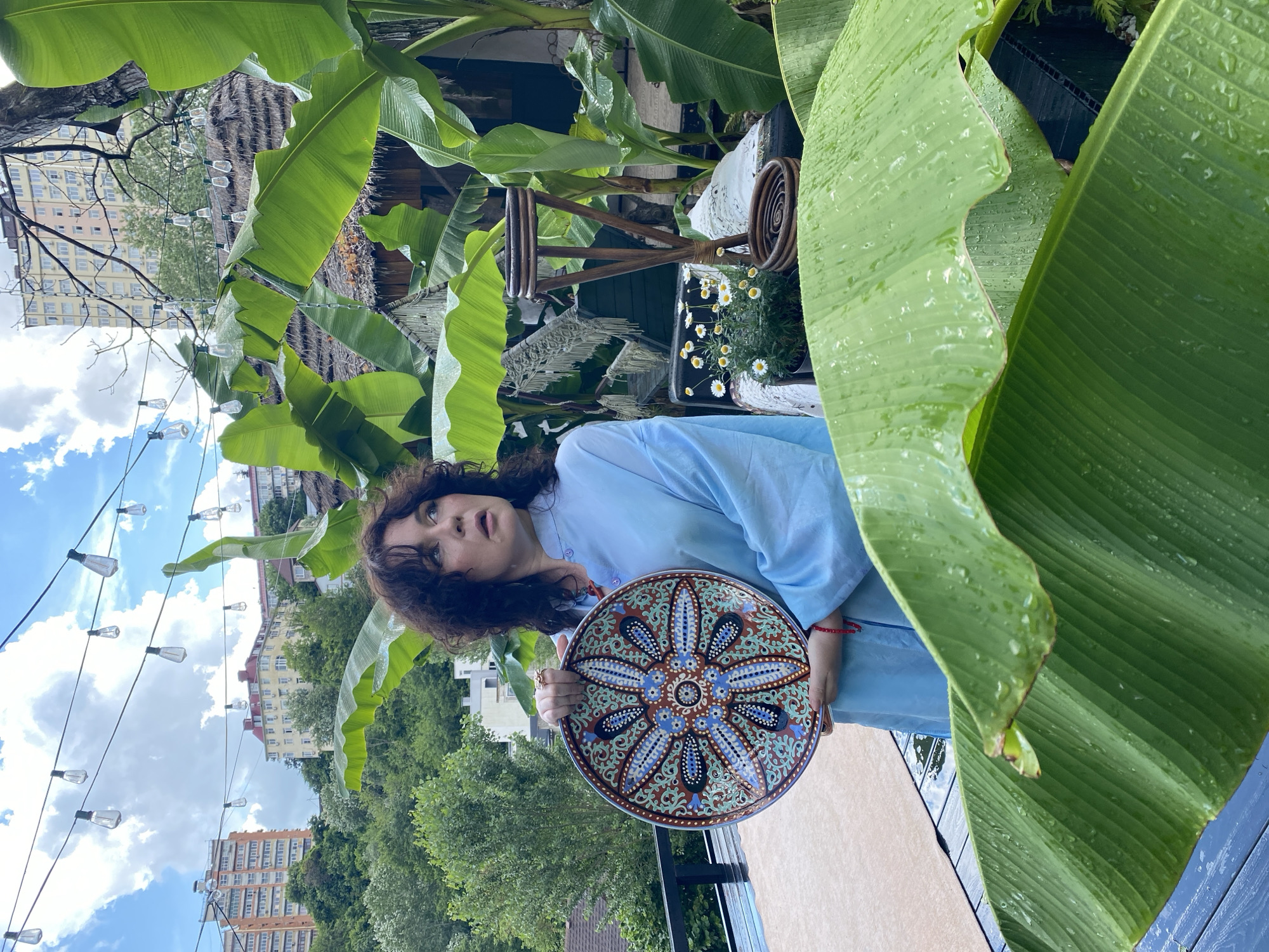 A woman stands on a rooftop terrace in Sochi, Russia, holding a circular decorative plate. She wears a light blue tunic and has dark, curly hair. The plate features a colorful, intricate floral pattern. The terrace is adorned with lush, large green banana leaves, some with water droplets on them, indicating recent rain. In the background, buildings are visible on a hillside, along with string lights overhead and a thatched roof structure. The sky is partly cloudy.