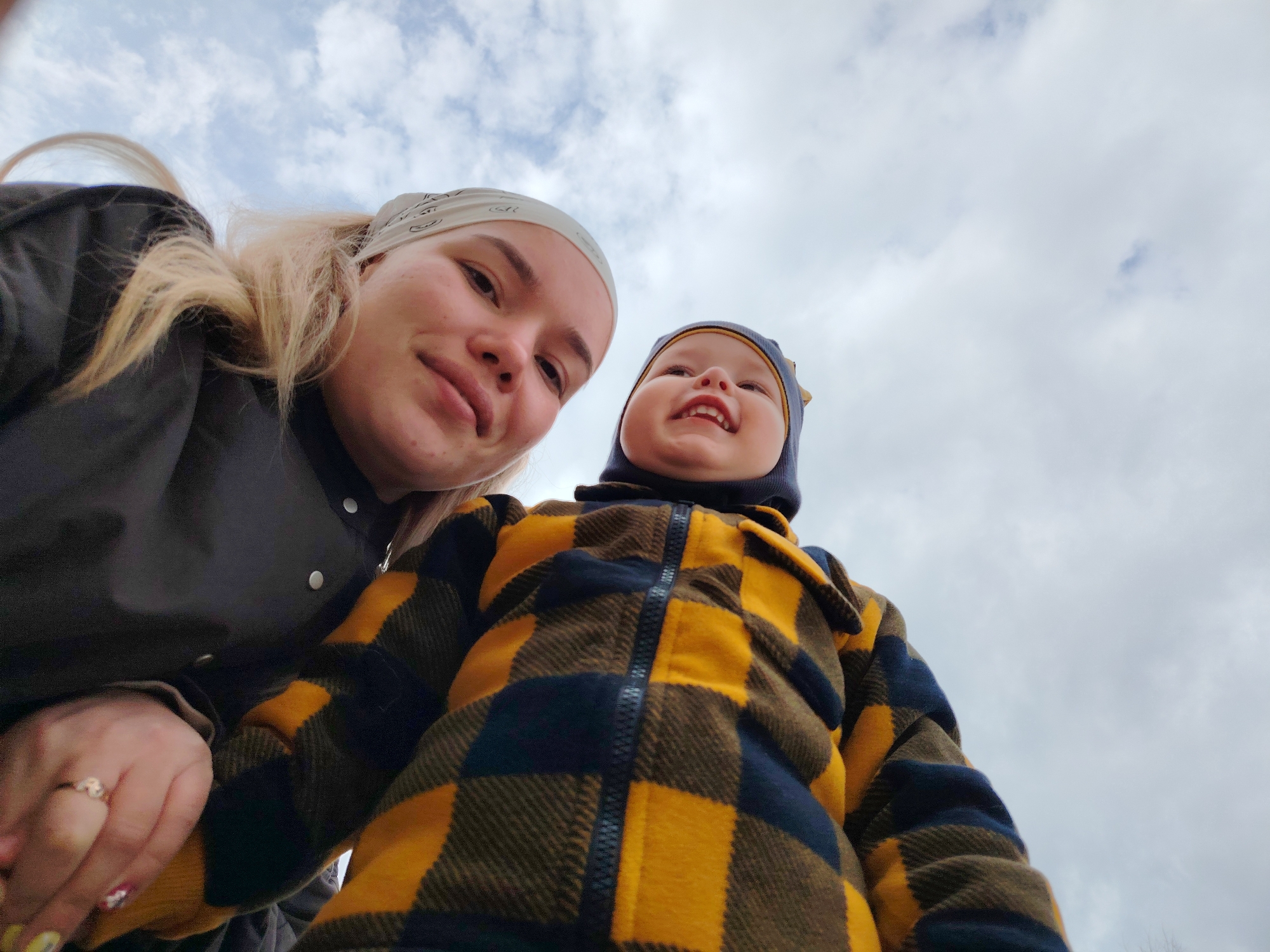 Two people, one adult female and one toddler, are positioned closely together, facing upwards towards the camera. The female is wearing a white headscarf and a dark jacket. The toddler is wearing a dark-colored hat and a yellow and brown plaid jacket. The sky is overcast with scattered clouds. The location is Markovskii, Russia.