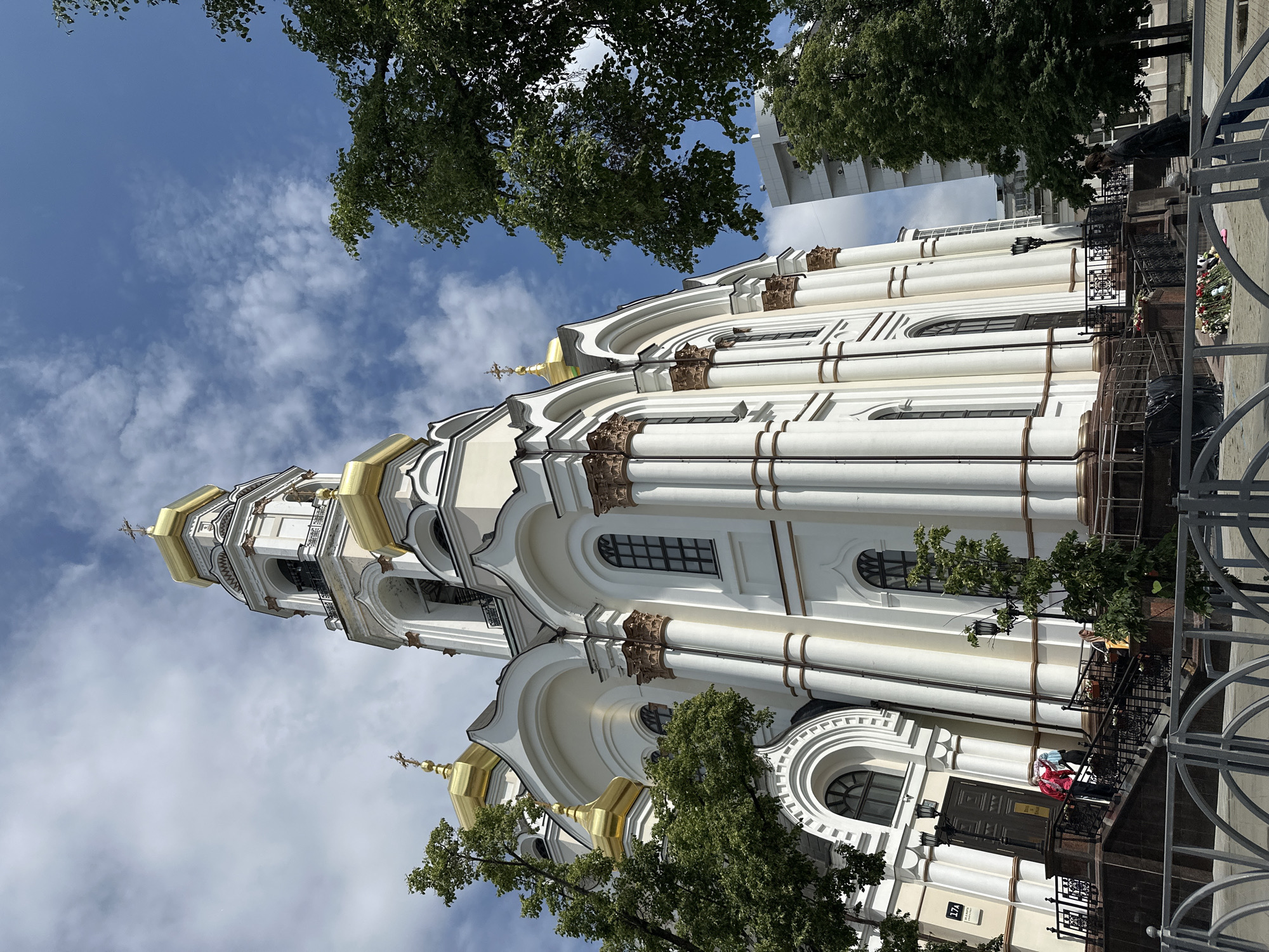 This is a low-angle, eye-level outdoor photograph of the Church of the Holy Trinity in Yekaterinburg, Russia. The church is predominantly white with gold domes, featuring classical architectural elements such as columns and arched windows. A tall bell tower with a gold dome and a cross rises on the right side of the church. Lush green trees frame the left and right sides of the church, and a modern, white building is visible behind the trees on the right. The sky is partly cloudy with patches of blue. In the foreground, a dark metal fence separates the viewer from the church. A set of stairs leads up to the church entrance, and several people are visible on the stairs and near the entrance. Two people are on the stairs, one wearing a red jacket, and another person in a dark coat is standing on the sidewalk to the right, looking towards the church. A cluster of flowers is placed on the ground in front of the fence.