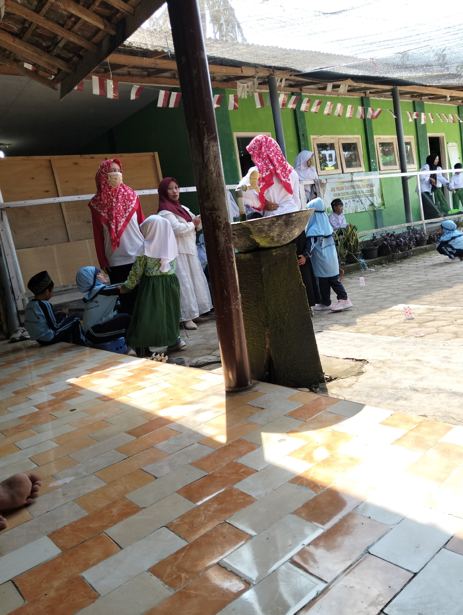 In Desa Wringin Putih, Indonesia, a group of individuals, primarily young girls in hijabs and uniforms, are gathered outdoors near a school building. The scene unfolds in an open area with a tiled floor in the foreground. Sunlight casts strong shadows, indicating a bright day.

Several girls, dressed in light blue and green uniforms with hijabs, are the central figures. One girl in a green skirt and a white top with a light blue hijab is looking down. Another girl in a light blue uniform and hijab is standing next to her. A woman wearing a red and white patterned hijab and a white top is also visible, standing further back. In the background, the green building of what appears to be a school or institution is visible, with windows and a sign. Flags, likely Indonesian flags given the colors, are strung along the top of the structure. Two young boys in blue uniforms and black peci caps are seated on the ground in the foreground, facing away from the camera. There are also a few other people in the background, including a woman in a dark hijab and white clothing, and another person in a white uniform. Small pink and white objects are scattered on the ground. The environment suggests a school or community gathering in a rural or semi-rural setting.