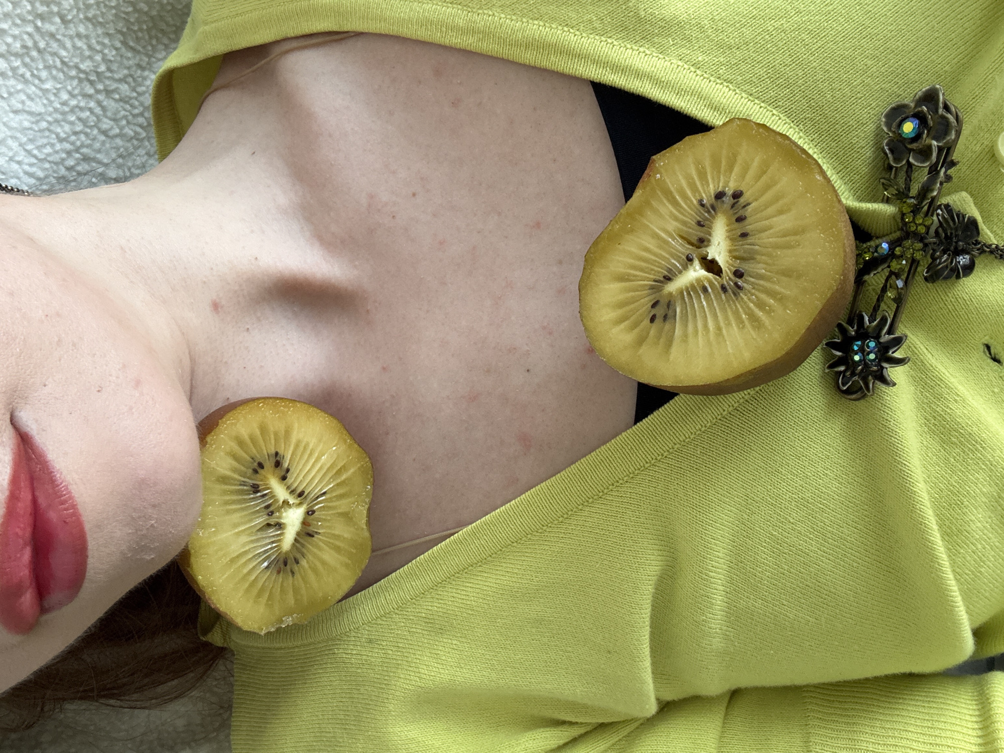 A person lies on a surface, with a partial view of their face showing red lipstick, and hair. The person is wearing a chartreuse sweater, adorned with a floral brooch. Two slices of golden kiwi are positioned on the person’s body. The scene is indoors, no location in Moscow, Russia, is evident.