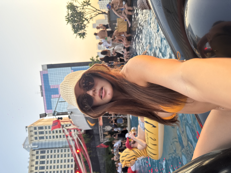 A young Caucasian female is depicted in a close-up, taking a selfie at a rooftop pool party during golden hour in Bangkok, Thailand. The scene is situated overlooking the distinctive Central Embassy complex in the Pathum Wan district, identifiable by its large pink/purple "T" shaped facade on a high-rise building in the background.
The primary subject, facing the camera, wears a light-colored woven bucket hat and dark sunglasses. Her long brown hair extends over her shoulders, and she wears an orange bikini top. She is seated partially submerged in the pool or on a black inflatable structure.
In the midground, a male subject with a beard, wearing a red baseball cap and sunglasses, reclines on a yellow tiger-shaped inflatable in the pool, holding a beverage. Numerous other individuals are present in and around the pool, some seated on the edge, some standing, engaged in social interaction.
The background features additional high-rise buildings and an event area on a lower terrace or platform adorned with red and white draped banners and a black flag, possibly an Albanian flag. The sky transitions from light blue to a warm orange hue. Small red specks are visible in the upper left portion of the sky, consistent with fallen confetti. A small tree is visible on the right side of the frame.