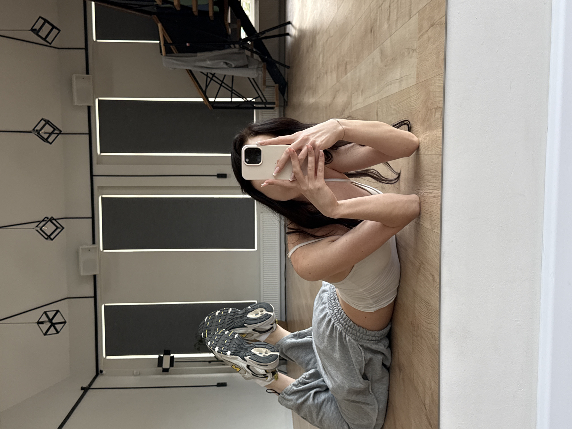 A young woman with dark hair is captured in a reflection, posing on a wooden floor in what appears to be a studio or home gym. She is wearing a white crop top and grey sweatpants, with athletic shoes on her feet. The woman is holding a smartphone and taking a selfie, with her face partially obscured by the device. Her pose is flexible and athletic, with her body contorted in a way that suggests she is engaging in a yoga or flexibility exercise.
The background visible in the reflection shows a clean, modern space with large windows or screens, some with black blinds. There are also geometric light fixtures hanging from the ceiling and a white speaker mounted on the wall. The overall setting suggests a well-lit, contemporary environment. The image is taken from a low angle, emphasizing the woman's pose and the reflective surface. No specific time of day or weather conditions are discernible. The woman's expression is not fully visible, but her posture conveys a sense of focus and athleticism. There is no visible text in the image.