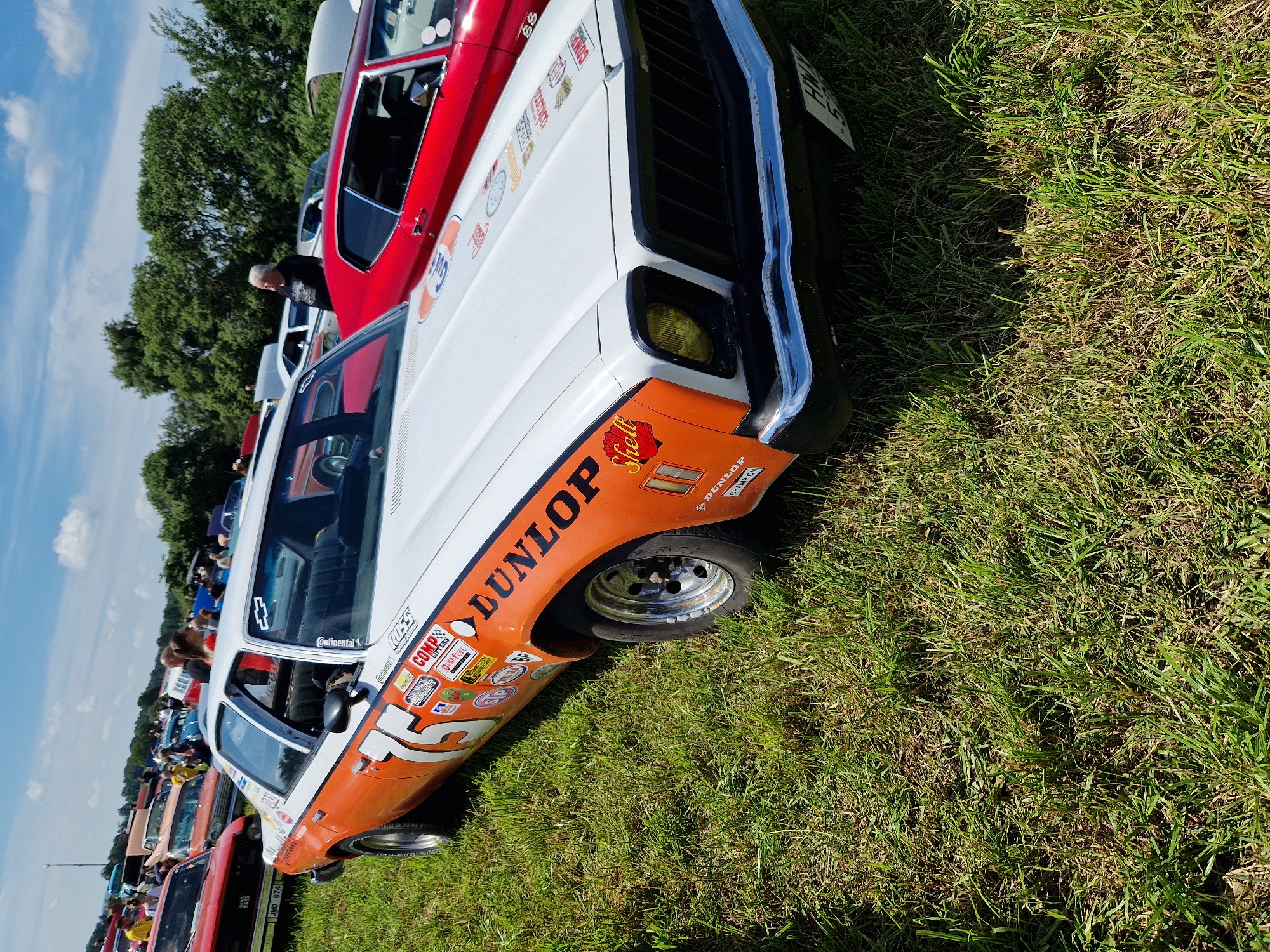 An exterior, eye-level, medium shot captures a classic, white and orange Chevrolet Nova coupe, race car, parked on a grassy field under a partly cloudy, blue sky. The car, with the number 75 emblazoned on its side, is covered in sponsor decals from brands like Dunlop, Shell, and STP. Its black grille and chrome bumper are visible, as is a yellow headlight. Behind the Nova, several other cars are parked, including a red coupe. The grass is green and appears recently mowed. The background features trees and a few people milling about, suggesting an outdoor car show or event. The overall setting is rural, with no identifiable city buildings or landmarks visible, but the presence of many cars and people indicates a gathering.