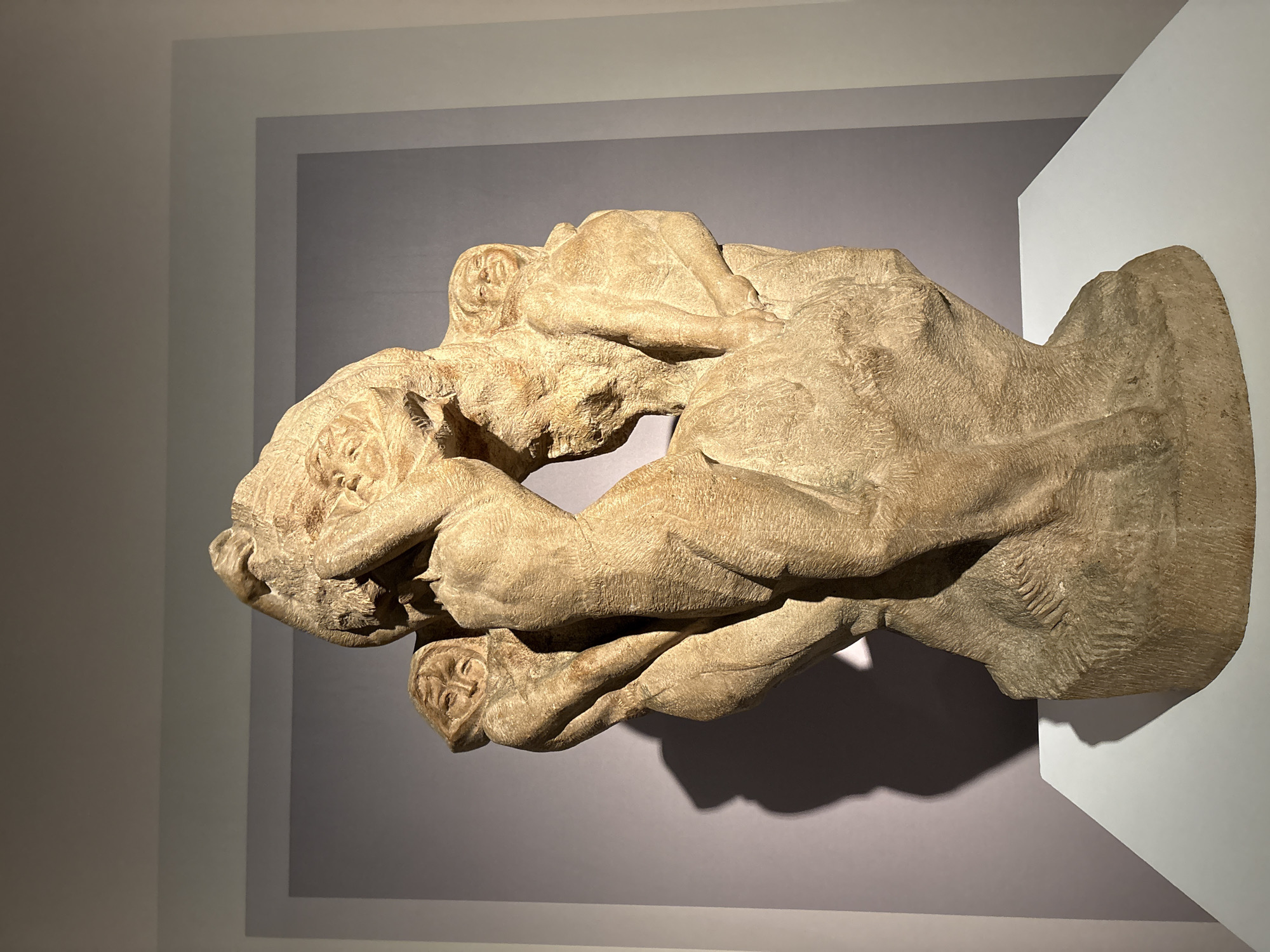 A stone sculpture, possibly a relief or a multi-figure composition, stands on a white pedestal in a dimly lit exhibition space. The sculpture depicts several figures intertwined, with a central, larger figure seemingly supporting or interacting with smaller figures clustered around it. The style appears somewhat rough-hewn and expressive, with visible chisel marks. The lighting casts strong shadows on the wall behind the sculpture, adding to the dramatic effect. The background wall is a neutral gray, with a lighter gray rectangular border. The sculpture is the sole focus of the image. The location in Pudong, China, is not discernible from the visual information presented.