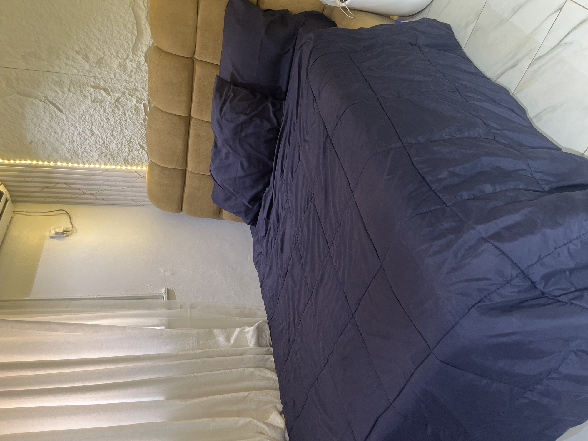 The image displays a bedroom with a focus on a neatly made bed. The bed is covered with a dark blue comforter that has a quilted diamond pattern. To the left of the bed, a tan-colored headboard is visible, composed of multiple square tufted sections. The wall behind the headboard has a textured, light-colored finish, possibly stone or a similar material.
Along the bottom of the image, sheer white curtains are drawn, with subtle horizontal folds that catch the light. Above the curtains, a string of small, warm-toned LED lights is illuminating the wall, suggesting it might be evening or a dimly lit interior. To the far left of the image, a white electrical outlet is mounted on a white wall.
The scene appears to be set indoors, likely in a residential bedroom. The overall impression is one of a tidy and perhaps modern living space. There are no people visible in the image, and no text is present. The lighting suggests a relaxed, indoor atmosphere. The location context of Abuja, Nigeria, provides a geographical setting for this domestic scene.