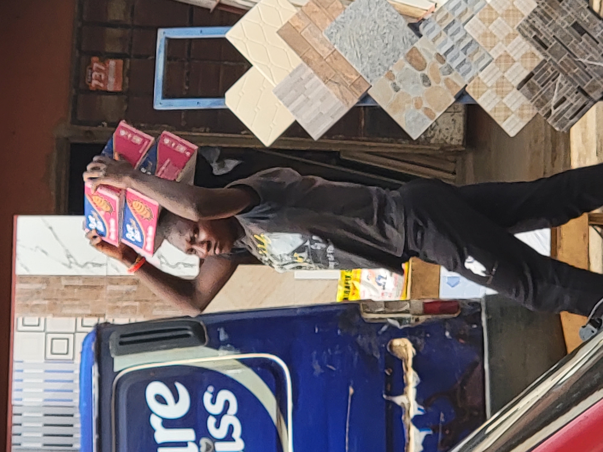 In Maiburiji, Nigeria, a person is carrying a stack of packaged goods on their head. They stand next to a blue vehicle with white lettering. Behind them, there's a display of various tile samples and a building facade.