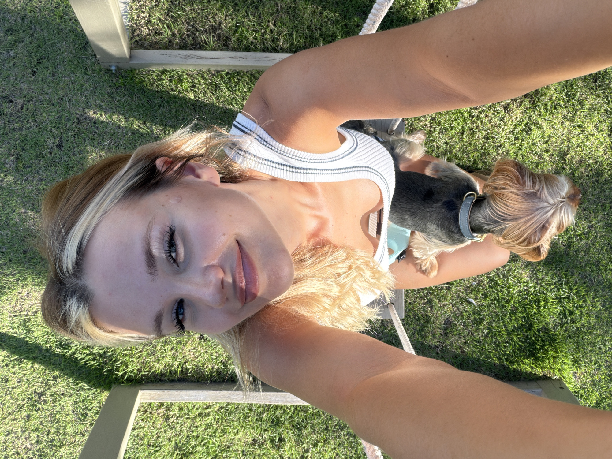 A young woman with blonde hair is taking a selfie outdoors with her small dog. She is wearing a white and grey striped tank top and a light blue sports bra or tank top underneath. Her dog, a Yorkshire Terrier, is sitting on her lap, facing away from the camera. The woman has on makeup, including mascara and lip gloss, and her eyes are open. The background is lush green grass, and part of a wooden structure is visible, possibly a bench or a table. The sunlight suggests it is daytime, likely morning or afternoon, and the lighting indicates clear weather. The woman appears relaxed and happy, enjoying time outdoors with her pet. The scene evokes a sense of leisure and connection with nature. There is no visible text in the image.