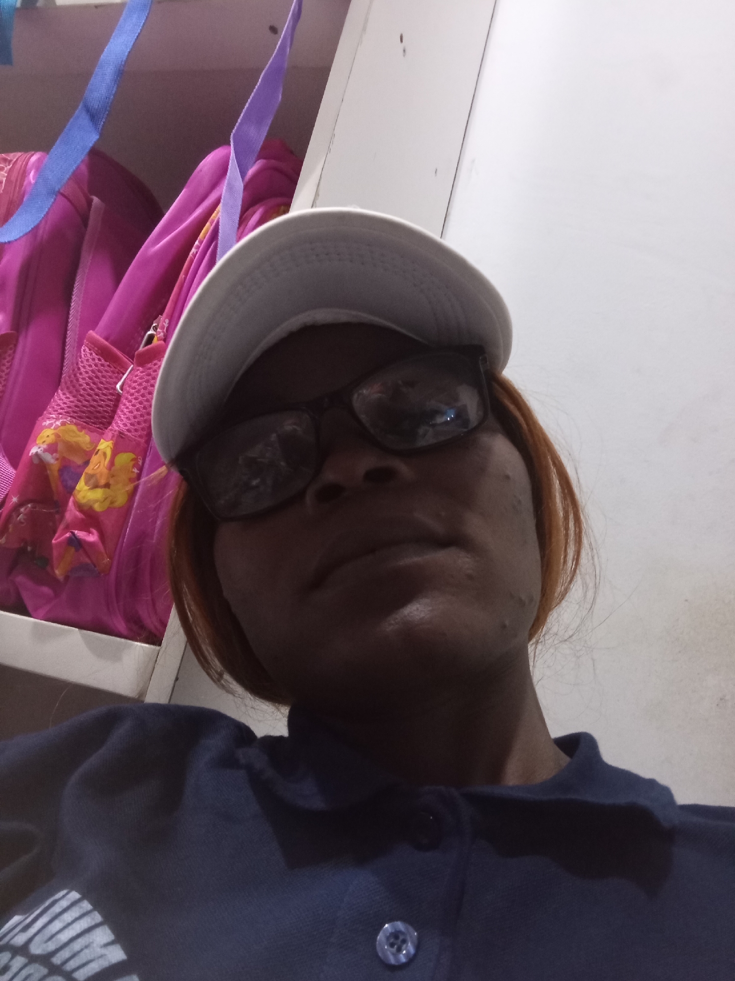 In the city of Howayi, Nigeria, a person wearing glasses and a white cap is seen in a close-up, low-angle selfie. They are wearing a dark blue collared shirt with buttons. Behind them, a pink backpack with cartoon characters is visible, along with blue and purple straps. The background appears to be an indoor setting, possibly a wall or shelf.