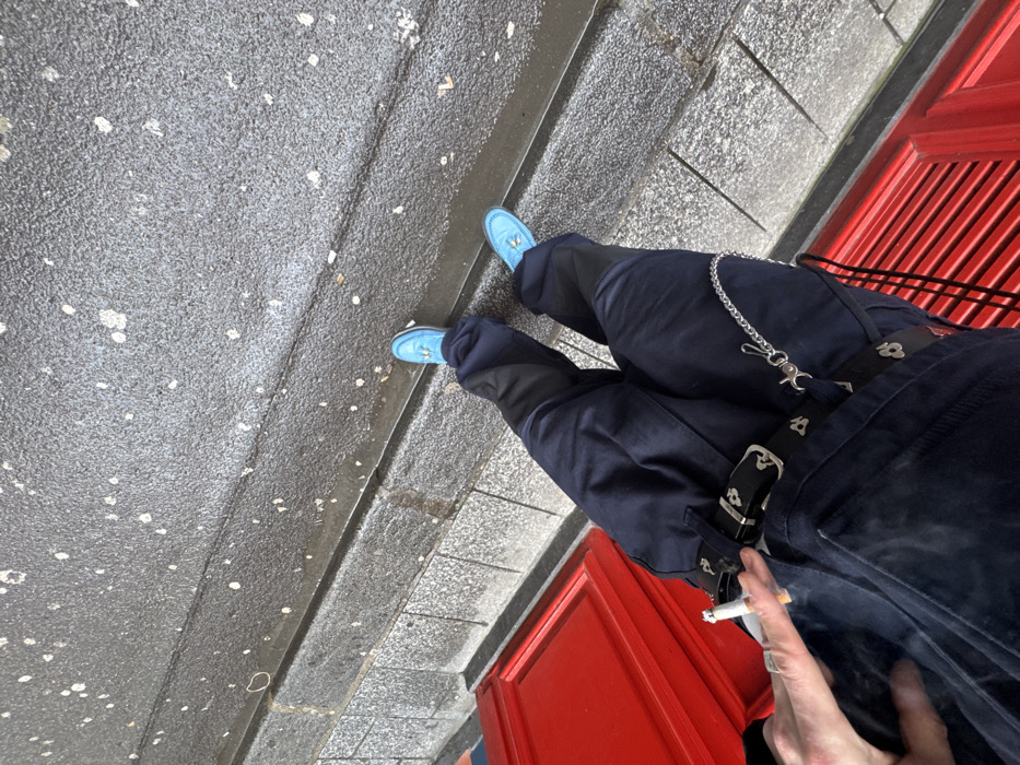 A person wearing navy blue pants, a black belt with white patterns, and light blue loafers is standing on gray steps next to a red doorway. The pants have a silver chain attached to them. The person is holding a lit cigarette in their hand. The scene is an overhead shot tilted to the right.