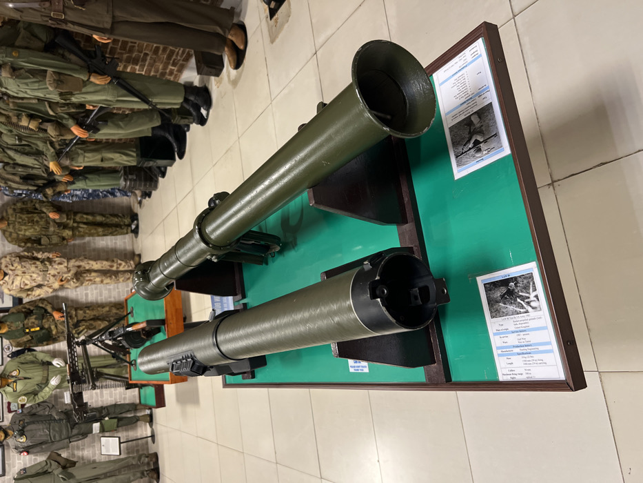 A museum exhibit features two green-painted rocket launchers mounted on a green background within a brown wooden frame. The launchers are displayed with information cards next to them. Mannequins dressed in various military uniforms stand to the left of the exhibit. The floor is light-colored tile.