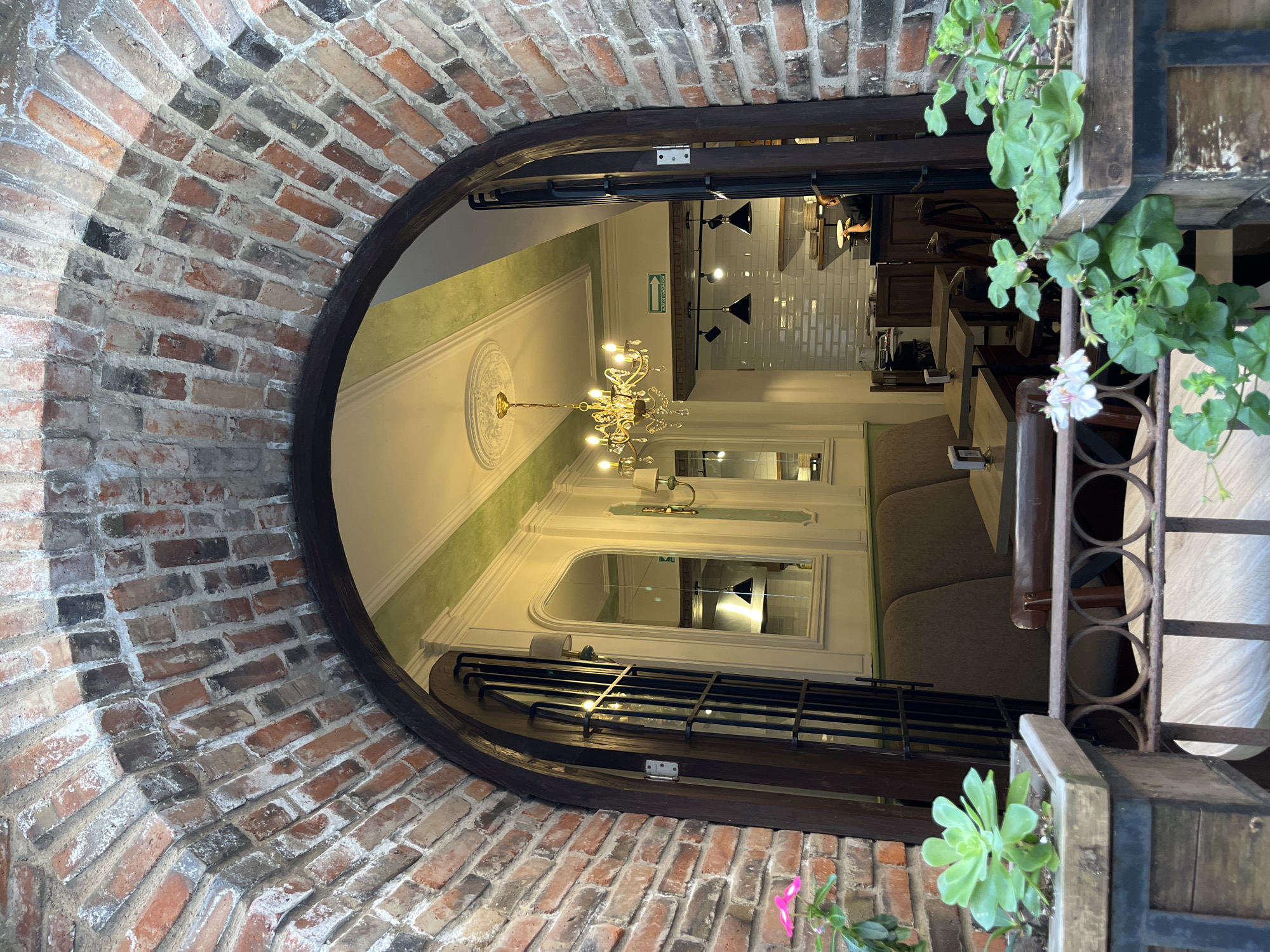The image shows an arched brick entryway to a building in what could be Domingo Arenas, Mexico, although it is impossible to confirm that from the image alone. The archway is made of red and some darkened bricks and has a dark wooden frame. Through the archway, the building's interior is visible, featuring a chandelier and interior décor with green and white color schemes. Furniture, including tables and chairs, suggests the space is likely a restaurant. The view through the arch is oriented to make the viewer feel disoriented. A planter with succulents and flowers is visible on the right side of the archway.