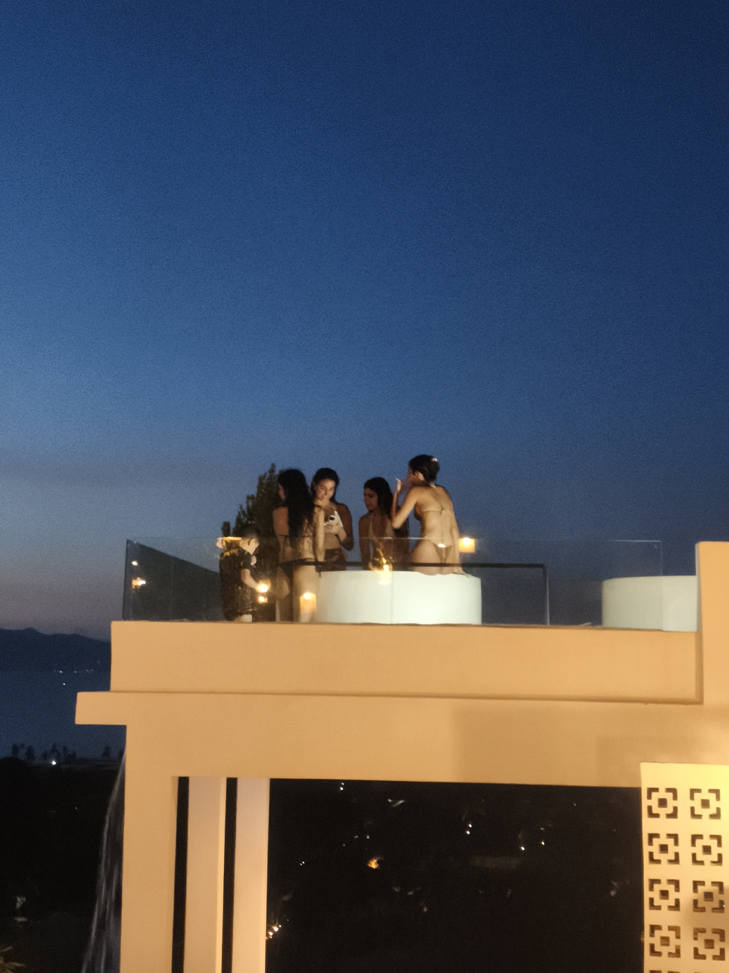 A group of four women are gathered on a rooftop patio at dusk, standing around a white, circular hot tub. The sky is a deep, clear blue, transitioning to a lighter hue near the horizon where mountains are visible in the distance. The women are wearing bikinis and appear to be engaged in conversation and leisure. One woman in the foreground is looking at her phone, while another stands next to her, looking in the same direction. The patio has a glass railing overlooking a city with lights visible in the darkening landscape. A patterned decorative panel is visible on the right side of the image, suggesting a modern architectural style. The scene is illuminated by ambient light from the sky and some artificial lighting, including candles placed on the edge of the hot tub. The location is identified as Baan Bang Rak, Thailand.