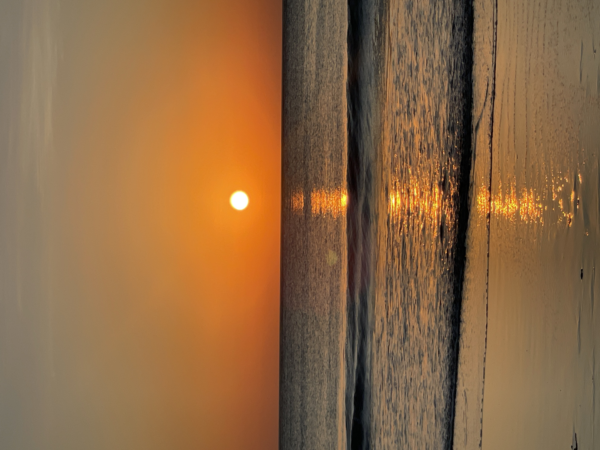A vertical, eye-level shot captures a tranquil seascape at sunset. The sky transitions from a pale gray at the top to a vibrant, deep orange as it meets the horizon. A bright, white sun, nearly fully set, is positioned centrally in the sky, casting a shimmering reflection that stretches across the water towards the viewer. The ocean is calm, with gentle waves lapping at the shore. The wet sand in the foreground reflects the warm hues of the sunset, creating a mirror-like effect. No people or animals are visible in the frame. The scene conveys a sense of peace and natural beauty. The location is Unknown, Unknown.