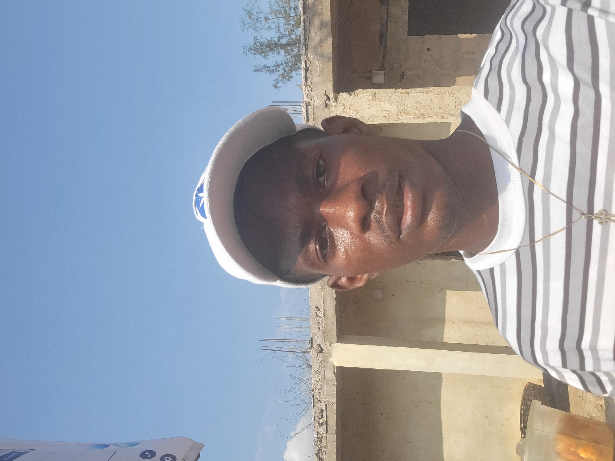 In Egbagu, Nigeria, a man is captured in a selfie, his face partially obscured by a white baseball cap with a blue logo. He is wearing a horizontally striped t-shirt in shades of gray and white, with a thin gold chain and a cross pendant visible. The background reveals an unfinished building with a concrete ledge adorned with exposed rebar and thorny branches, under a clear blue sky. To the left, a clear plastic cup filled with orange-colored food items sits next to a woven basket.