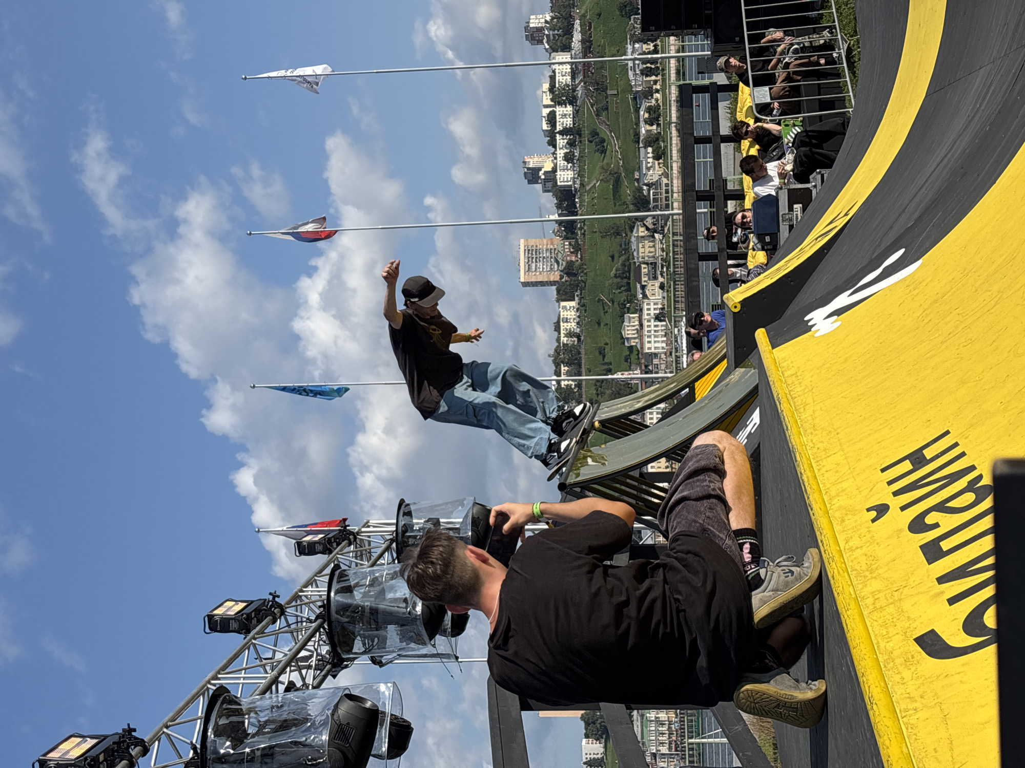 A male skateboarder in mid-air performs a trick on a yellow and black ramp with a cityscape and river in the background. The skateboarder wears a black t-shirt, blue jeans, a baseball cap, and sneakers. In the foreground, a man with a black t-shirt and shorts kneels, holding a camera to film the skateboarder. He has short brown hair and is wearing sneakers and socks. The event appears to be outdoors, with a clear blue sky and scattered clouds. Flags are visible in the background, including Russian flags, and large lighting rigs are positioned on a metal structure. The cityscape shows buildings of varying heights situated on a hill overlooking the river. The word "билайН" is visible in large black lettering on the yellow ramp. A crowd of spectators is seated behind a fence on the right side of the image, watching the event. The location is the Nizhny Novgorod Riverfront, Russia.