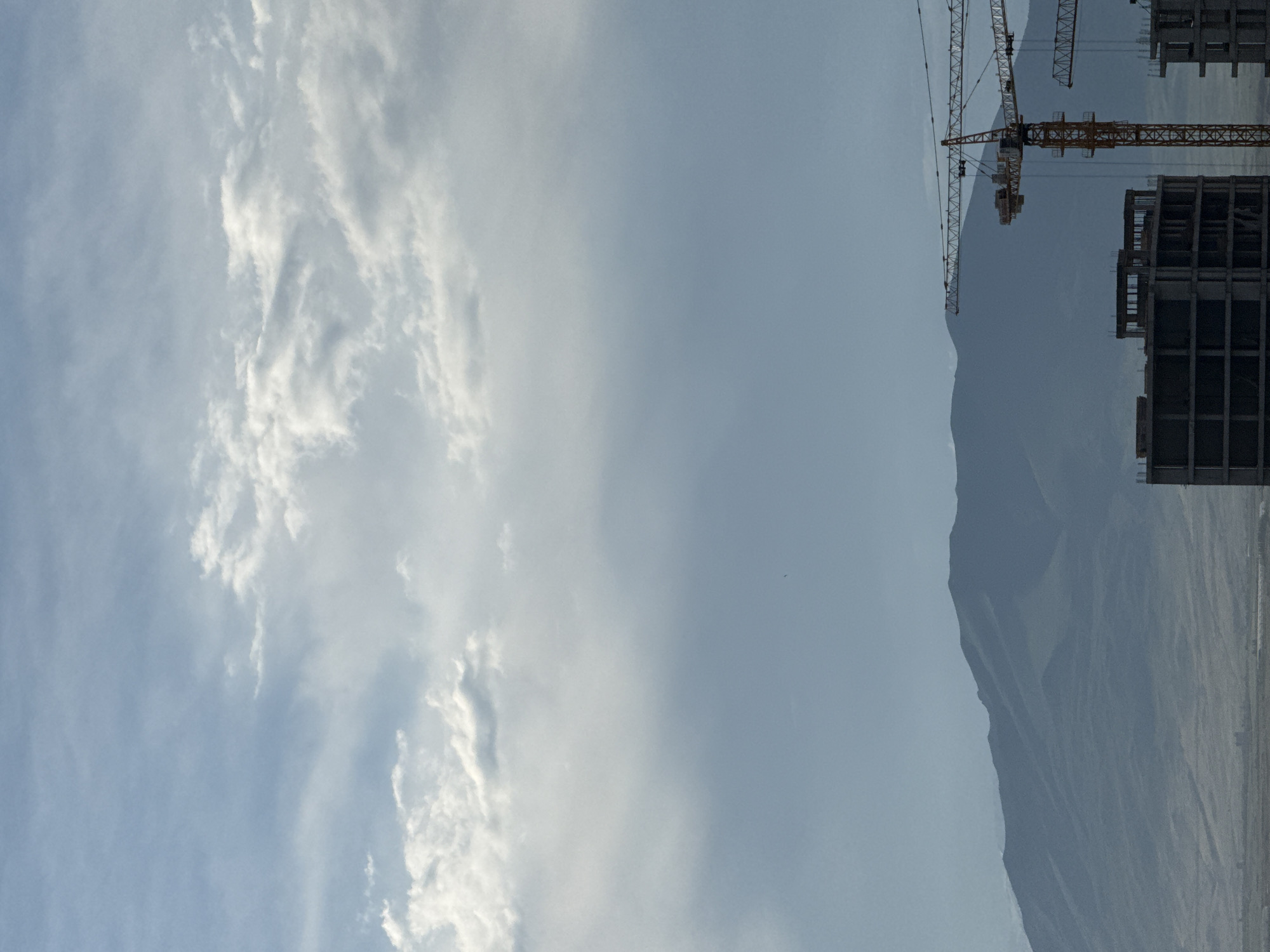 The image depicts a construction site in Abovyan, Armenia, with mountains in the background. In the foreground, there are two partially constructed buildings, one of which is a concrete frame structure with multiple floors. Yellow construction cranes are present, with one prominent crane positioned between the buildings. The sky is partially cloudy, with soft white clouds against a pale blue backdrop. A small, dark bird is visible in the sky, adding a touch of life to the scene. The overall atmosphere is one of urban development against a natural mountainous landscape.