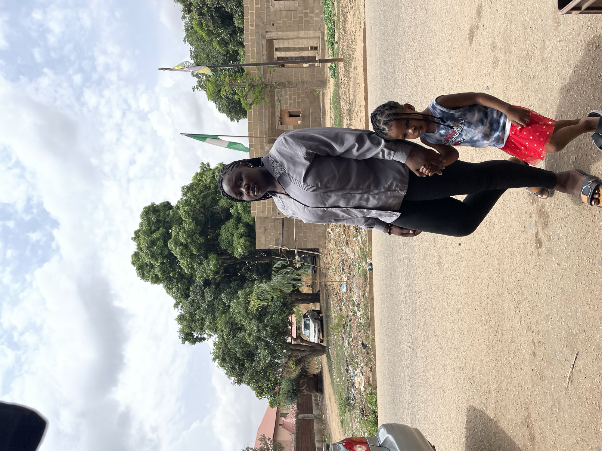 A full-length outdoor shot shows two people holding hands on a paved road in Jos, Nigeria. An adult female with braided hair stands close to a child, also with braided hair. The adult is wearing a gray, long-sleeved shirt and black leggings. The child is wearing a gray-blue patterned tank top and a short red skirt with white stars. Both are facing the camera at a slight angle. The background includes a building under construction made of gray bricks, green trees, and a Nigerian flag. Litter is visible on the side of the road. Part of a silver car is visible in the lower right corner. The sky is cloudy.