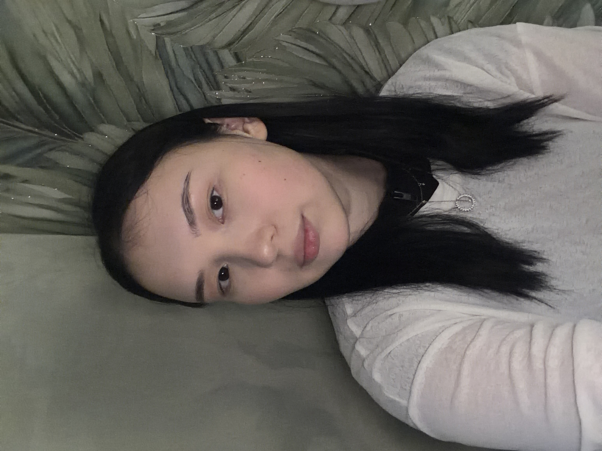 A young woman with long, dark hair lies on her side, resting her head on a surface covered with a green, leaf-patterned fabric. She is wearing a white long-sleeved shirt. The lighting is soft, suggesting an indoor setting, possibly a home, during the day. Her face is turned towards the camera, and she has a neutral expression. A delicate silver necklace with a circular pendant is visible around her neck. The background behind her head shows a green, textured wall or fabric, possibly a decorative backdrop. The overall scene conveys a sense of relaxation and casualness. No text is visible in the image. The provided location context is Chaikovsky, Russia.