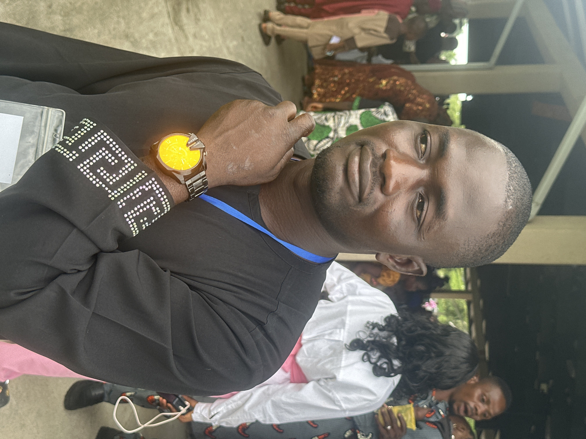 The image shows a man in the foreground, facing slightly to the left, with his hand raised to his chin. He is wearing a black, long-sleeved shirt with a blue lanyard around his neck. A prominent watch with a bright yellow face is visible on his left wrist. The cuff of his shirt is adorned with a pattern of small, white, diamond-shaped elements. In the background, other people are present, some out of focus, wearing colorful clothing. The setting appears to be an outdoor or semi-outdoor area, possibly a gathering or event. The architectural elements in the background suggest a building with columns and a roof. The overall scene conveys a sense of community or social interaction. The location is specified as Itoga, Nigeria.