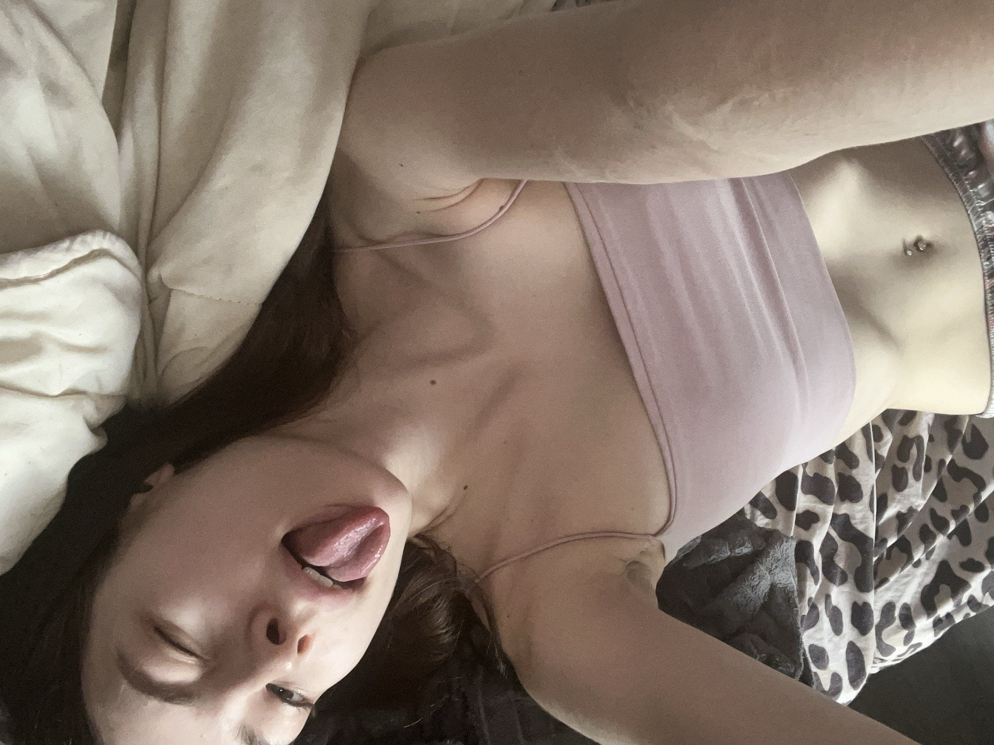 A young woman is lying on a bed with animal print sheets and a grey blanket. She is wearing a light pink tank top and silver pajama shorts. She is lying on her back and tilted her head to the side, sticking out her tongue and squinting. A white pillow is visible in the top left corner. Her stomach is visible with a belly button piercing. There is no indication of the location being Saint Petersburg, Russia.